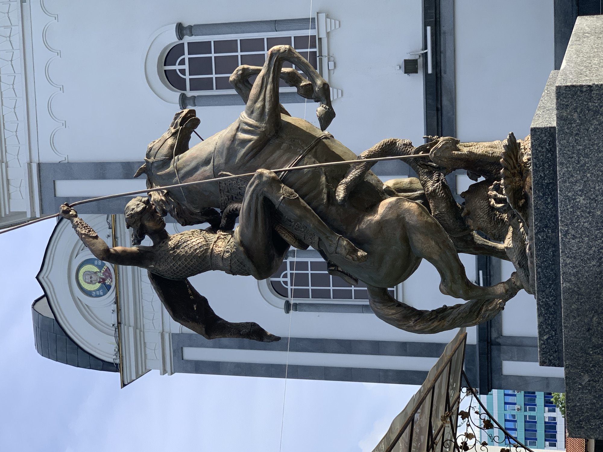 The image shows a bronze statue of Saint George slaying the dragon, with a church in the background. Saint George is depicted on horseback, thrusting a spear into the dragon's head. The church features white walls with gray accents and arched windows. The statue is positioned atop a dark granite base. A wrought iron fence with decorative vine details is visible in the lower part of the frame. In the distance, a building with blue and white windows can be seen. The statue is located in the city of Unknown, Unknown.