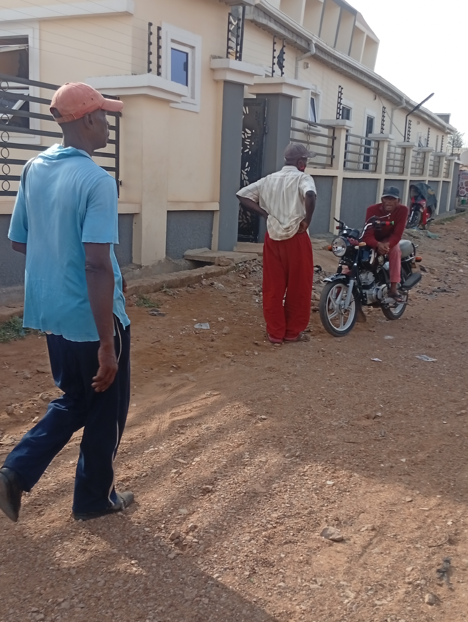 The image is captured in what appears to be Sabon Tasha, Nigeria. The setting is a dusty, unpaved road adjacent to a row of single-story residential buildings with decorative ironwork on the windows and security features on the walls.
Three individuals are in the foreground. On the left, a man wearing a blue t-shirt, dark sweatpants with a white stripe, and a peach-colored baseball cap is walking away from the camera. Further down the road, a man with a light-colored button-up shirt and red pants stands with his hands on his hips. A third man sits on a black motorcycle, wearing a red long-sleeved shirt and a dark cap. Other motorcycles are parked further down the road. The atmosphere appears to be daytime, with natural light. The road is covered with loose gravel and dirt.
