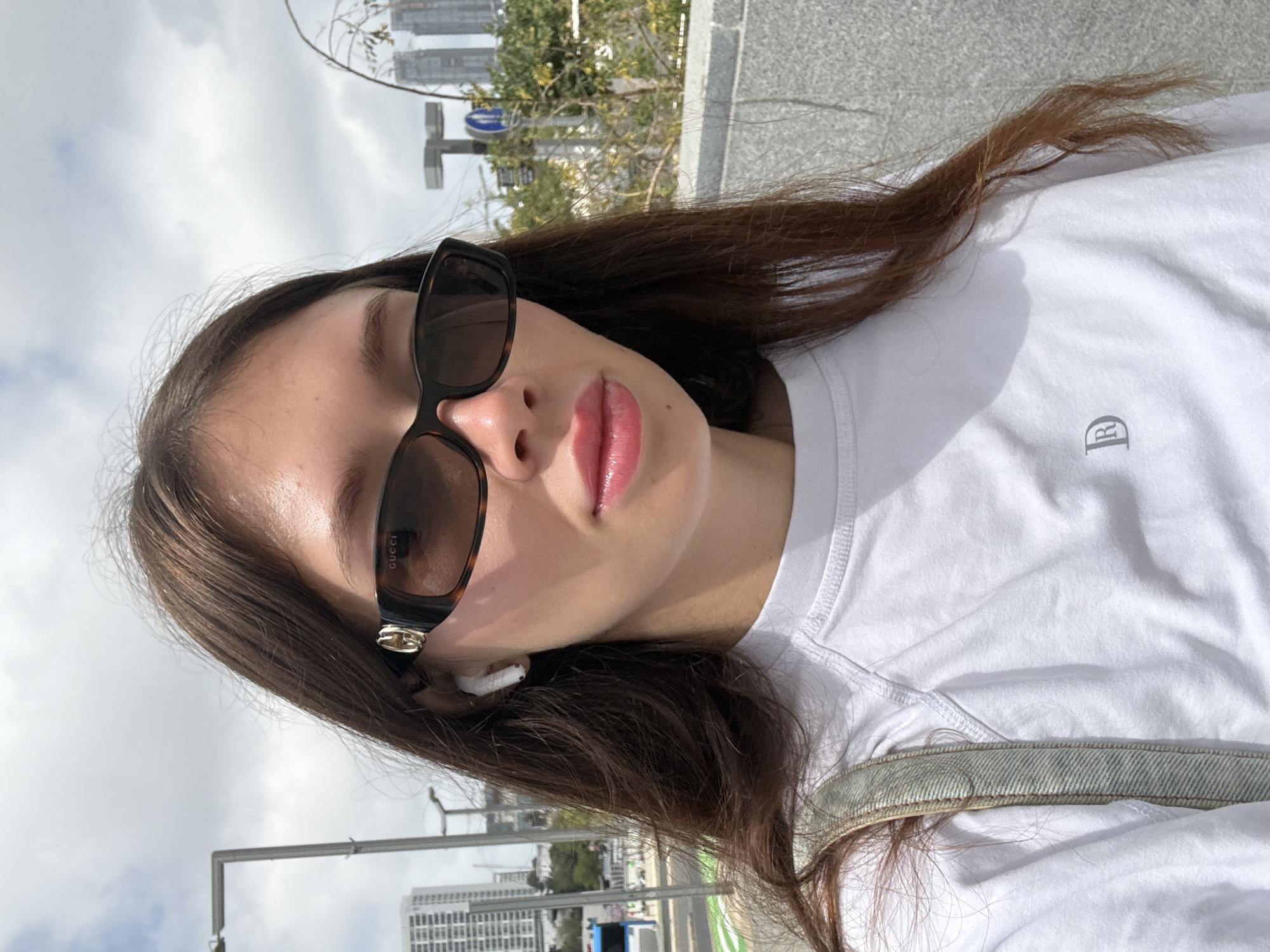 A woman is taking a selfie in Tel Aviv, Israel. She is wearing Gucci sunglasses and white AirPods. She has long brown hair, and she is wearing a white t-shirt. A denim jacket is visible. The backdrop is a city street with buildings in the distance.
