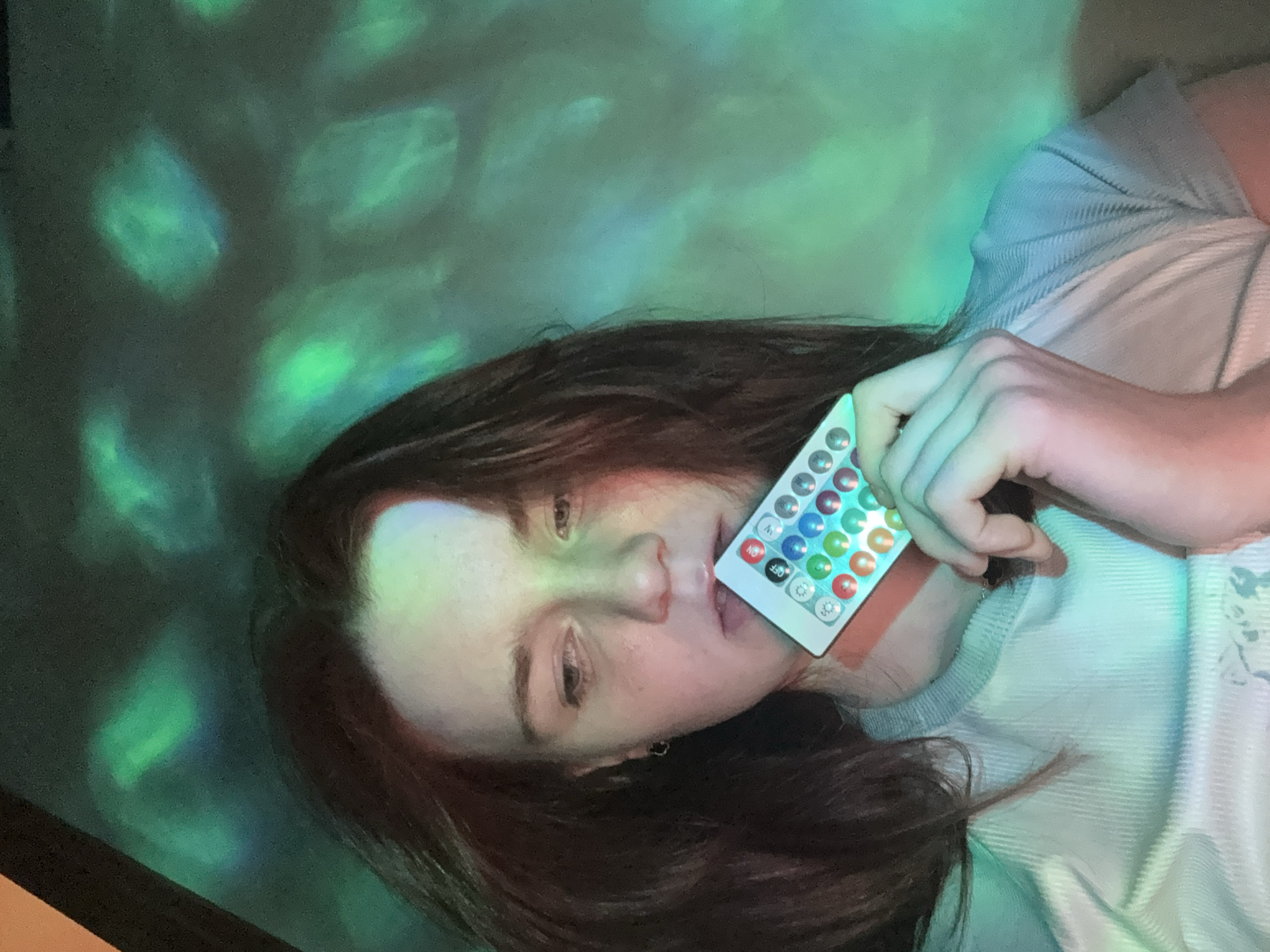 A young person with long, dark hair is depicted lying down, illuminated by an ethereal green and blue light. The image is taken from a slightly overhead perspective, with the person's body oriented diagonally across the frame.
The central subject, likely female, has fair skin and a calm, possibly contemplative expression, with her gaze directed slightly away from the camera. Her dark hair is spread around her head. She is wearing a light-colored, ribbed top or sweater.
Her right hand holds a small, white remote control with numerous colorful buttons, which she places near her mouth, almost touching her lips. The remote features rows of circular buttons in various colors including red, green, blue, and white, as well as several shades in between. Visible text and symbols on the remote include "ON," "OFF," "M," "S," and gear/settings icons.
The background is dominated by abstract, shifting patterns of green and blue light, suggesting an indoor setting with a light projector or LED lights in a dim environment, likely evening or night. These lights cast a greenish glow on the person's face, hand, and hair. There is also a subtle reddish/pinkish hue illuminating parts of her face and clothing from the right side. The overall mood is serene and somewhat dreamlike, with the unique lighting creating an immersive atmosphere. No specific location cues beyond the implied indoor setting are visible.