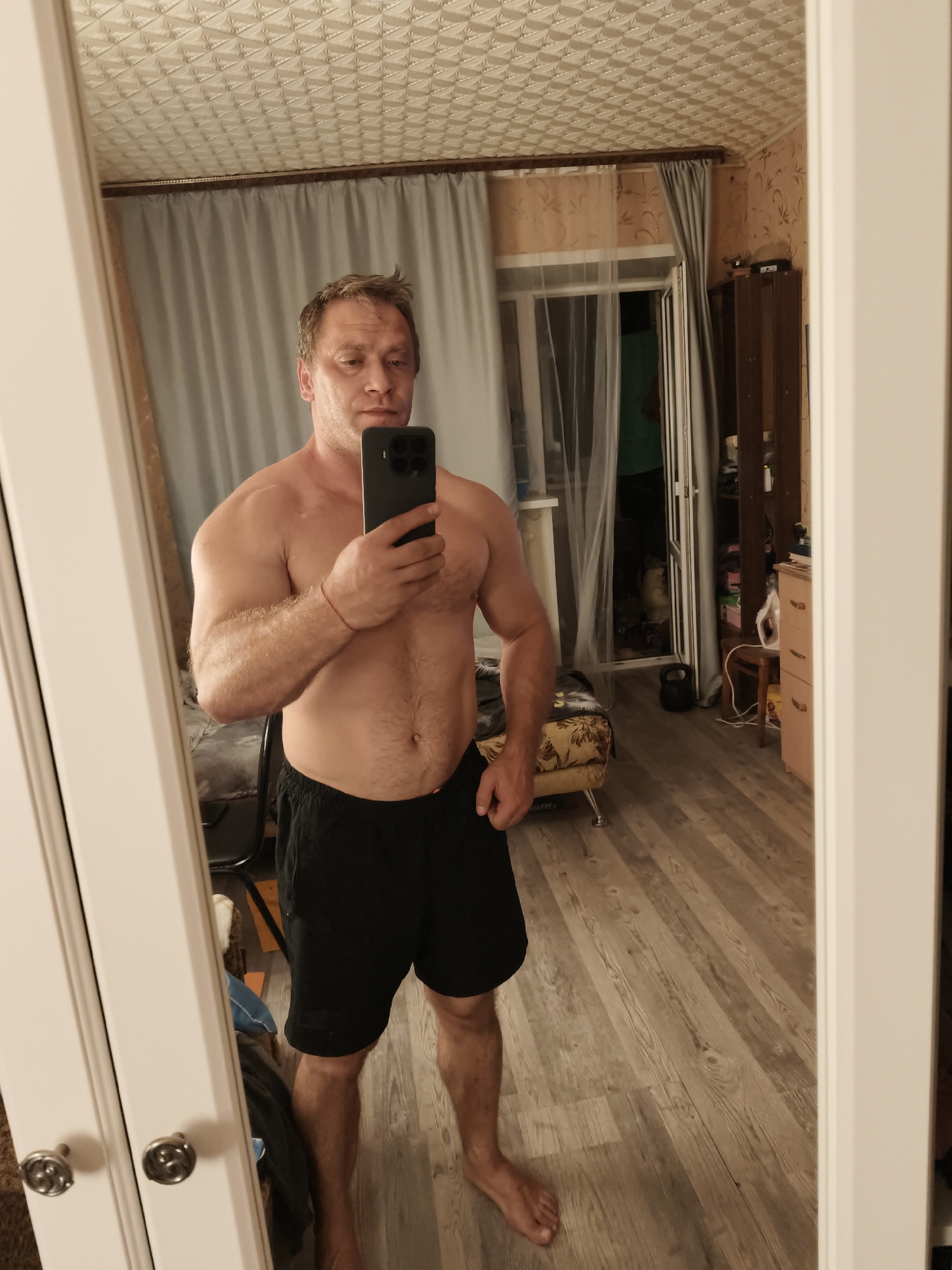 A shirtless man with light brown hair and a fair complexion is taking a mirror selfie in a room. He is wearing black athletic shorts. The man is standing in front of a full-length mirror, holding a smartphone in his right hand. The room appears to be a bedroom or living area, with wooden flooring, curtains, a dresser, and other household items visible in the reflection. The lighting suggests it is indoors during the day or evening. The specific location in Star, Russia cannot be determined from the image.