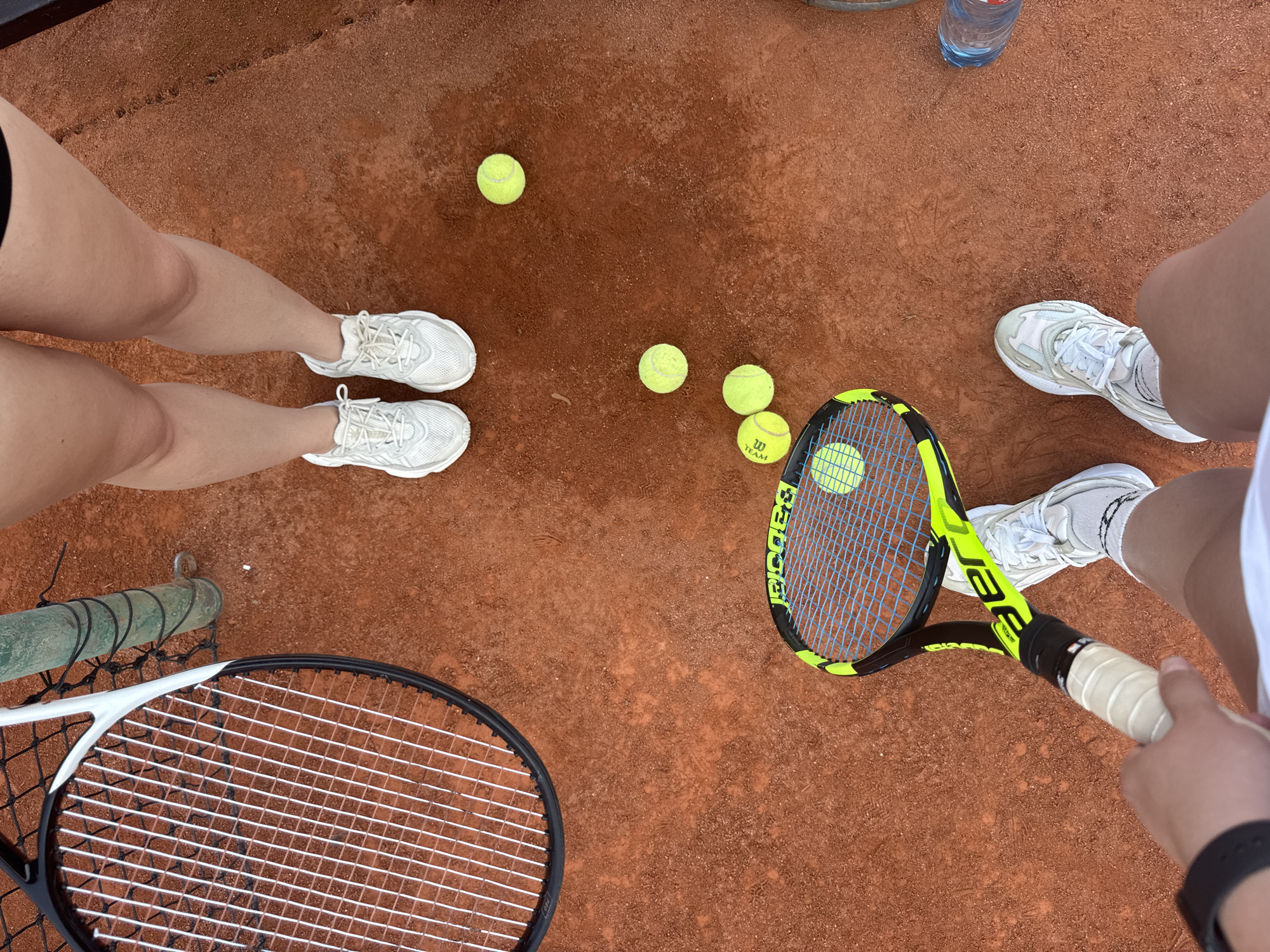 The image captures a top-down view of a tennis court, likely in Neubiberg, Germany. The court's surface is a reddish-brown clay, indicating an outdoor setting. Several tennis balls are scattered across the court, with at least five visible. Two tennis rackets are present, one with a neon green and black frame and blue strings, and another, partially visible, with a black frame and white strings.
The legs of at least two individuals are visible, wearing white tennis shoes and athletic shorts. One person's legs are on the left side of the frame, wearing white sneakers. Another person's legs are on the right, also wearing white sneakers and what appears to be a white skirt or shorts. One person's hand, wearing a black watch, is holding the handle of the neon green racket, suggesting they are actively playing or preparing to play.
A partially visible water bottle with a blue cap is also on the court. The lighting suggests it is daytime, and the image does not provide specific weather details, though the clay court appears slightly damp in places. The overall scene conveys a moment of preparation or a pause during a tennis match. Text visible on one of the tennis balls includes "W TEAM."