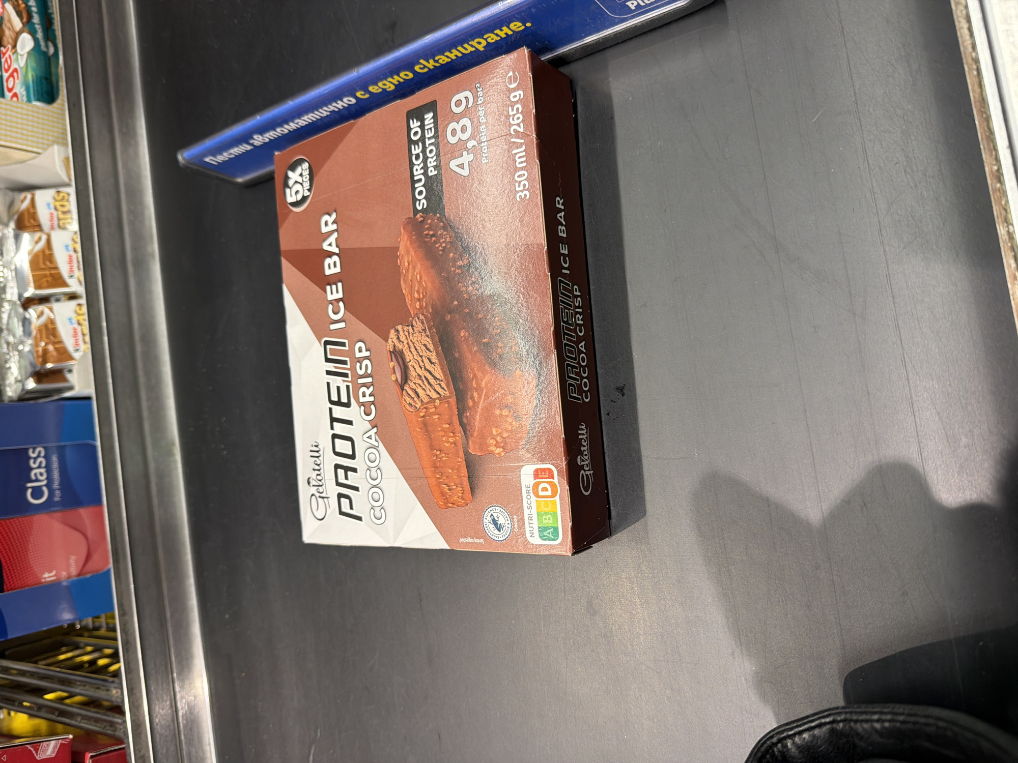The image shows a box of Gelatelli Protein Ice Bar Cocoa Crisp (5 pieces) on a supermarket checkout counter. Other items visible in the background include boxes of Kinder chocolates and a blue box of Class tissues. The text "Пести автоматично с едно сканиране." (Pesti automatichno s edno skanirane) is printed on a blue shelf. The "NUTRI-SCORE" is visible, which would indicate that this product is for sale in a European country. Given the Bulgarian text on the shelf, it would be accurate to assume that this is a supermarket in Sofia, Bulgaria.