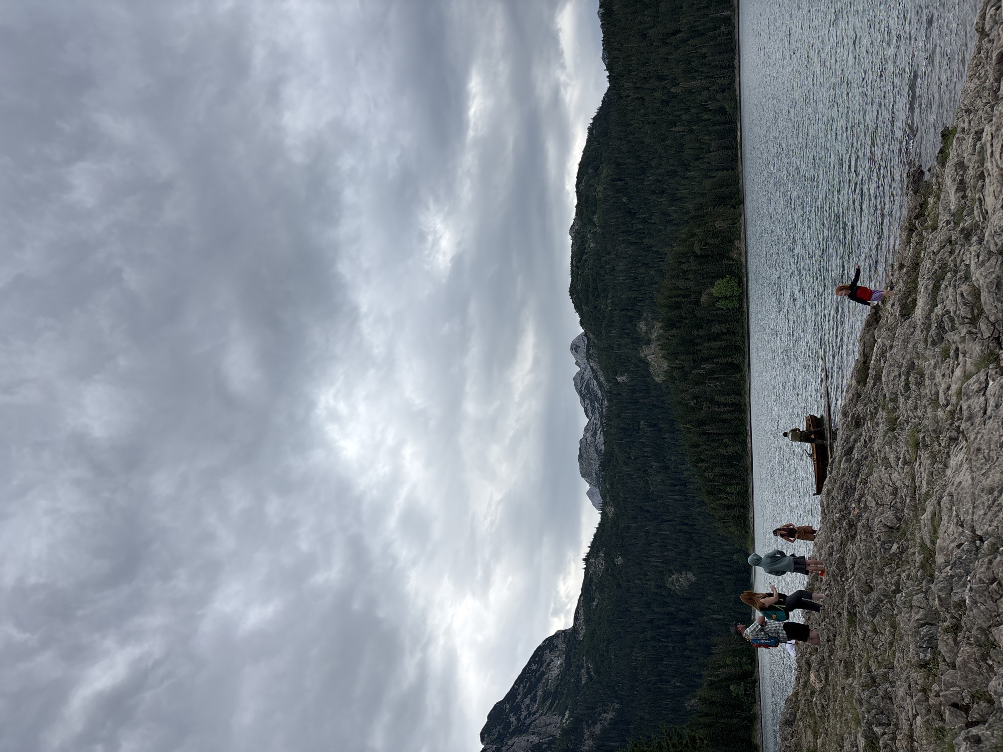 The image depicts a rocky shoreline of a lake in Pitomine, Montenegro. A group of five people are visible on the shore. Three individuals are standing together on the left side of the frame, one wearing a backpack and another holding a phone. Two more people are standing closer to the water on the right side. In the background, a small wooden boat is moored to a floating dock, with one person in or near it. The lake extends across the middle of the frame, and a dense, dark green forest covers the slopes of mountains that rise in the distance. The sky is overcast with heavy, gray clouds, suggesting a cool or potentially rainy day. The rocky terrain of the foreground is covered with various sizes of stones.
