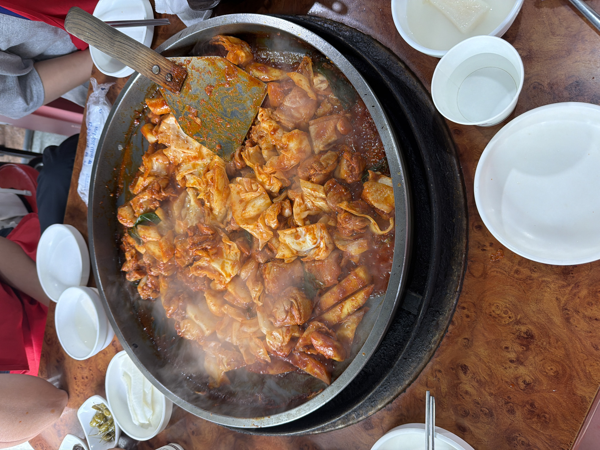 A close-up overhead shot captures a sizzling pan of dakgalbi, a popular Korean stir-fried chicken dish, served in a large, round, shallow metal pan on a wooden table. The dakgalbi, a vibrant mixture of chicken, cabbage, and a red, gochujang-based sauce, is actively steaming and bubbling, indicating it is freshly cooked. A metal spatula with a wooden handle is resting in the pan, partially obscuring the dish. Several small white bowls are arranged around the pan, containing side dishes such as pickled peppers and sliced radish. Two individuals, one wearing a red shirt and another a gray sweatshirt, are partially visible in the upper portion of the image, suggesting they are dining at the table. The table itself has a distinctive wood grain pattern. The setting appears to be a casual restaurant in Chuncheon-si, South Korea, a city renowned for its dakgalbi.
