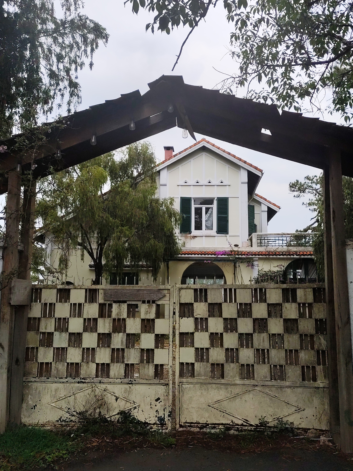 A weathered gate in Da Lat, Vietnam, frames a two-story house. The gate is painted in a checkerboard pattern with peeling white paint, a diamond design on its lower half, and encroaching weeds. Above, a wooden pergola with string lights provides a partial roof. The house features a red-tiled roof, green shutters on the upper windows, a balcony, and arched openings on the ground floor. Trees and foliage partially obscure the view of the house and overcast sky.