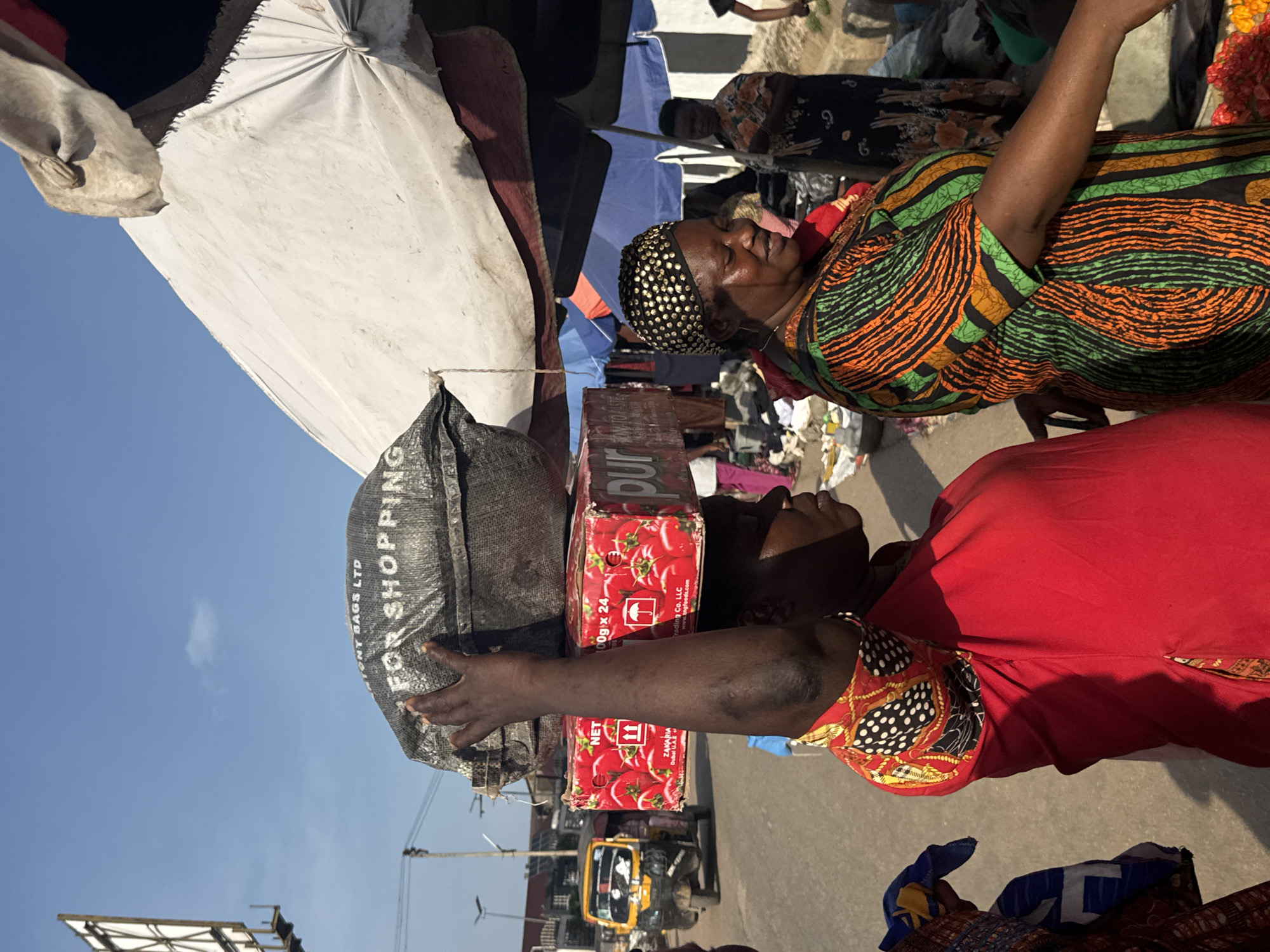 The image shows two women at what appears to be an outdoor market in Onitsha, Nigeria. The woman in the foreground, facing the camera and slightly to the right, is wearing a vibrant red dress and a headscarf with a black and white polka dot pattern. She has her arm extended, touching a stack of red boxes that are labeled with images of tomatoes and text that reads "NET 100g x 24", "Dur", and "Trading Co. LLC Dubai UAE". Beside the boxes is a large black bag with white lettering that says "ONY BAGS LTD FOR SHOPPING". The second woman is to the left of the foreground woman, wearing a colorful, patterned dress and a black headscarf adorned with gold embellishments. She is looking to her right and smiling. Behind them, there are tents or awnings, suggesting a market setting. The sky is a clear blue, indicating it is daytime. In the background, a yellow auto-rickshaw is visible on a street, and there are other market stalls and goods. The overall scene suggests a busy marketplace environment.