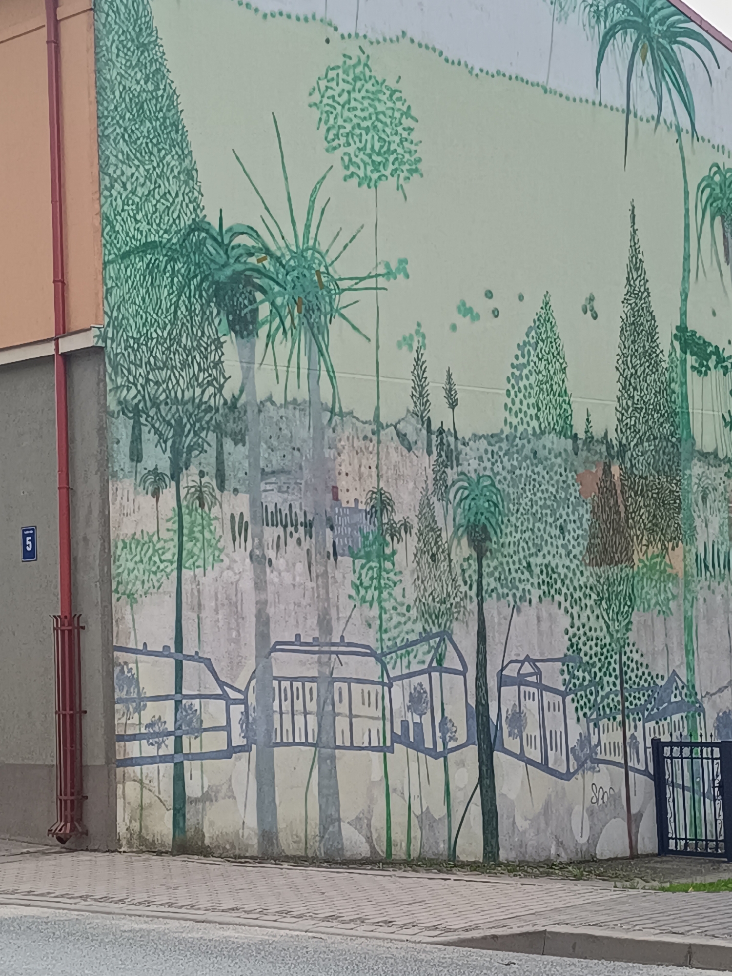 The image depicts a mural on the side of a building in Saldus, Latvia. The mural features a stylized landscape with various trees, including palm trees and densely packed greenery, depicted in shades of green. In the foreground, there are outlines of buildings and fences painted in gray and blue. A red pipe runs vertically along the left side of the building, adjacent to a small blue sign with the number "5". The building itself has a beige and gray facade. The mural is situated next to a sidewalk and a street.