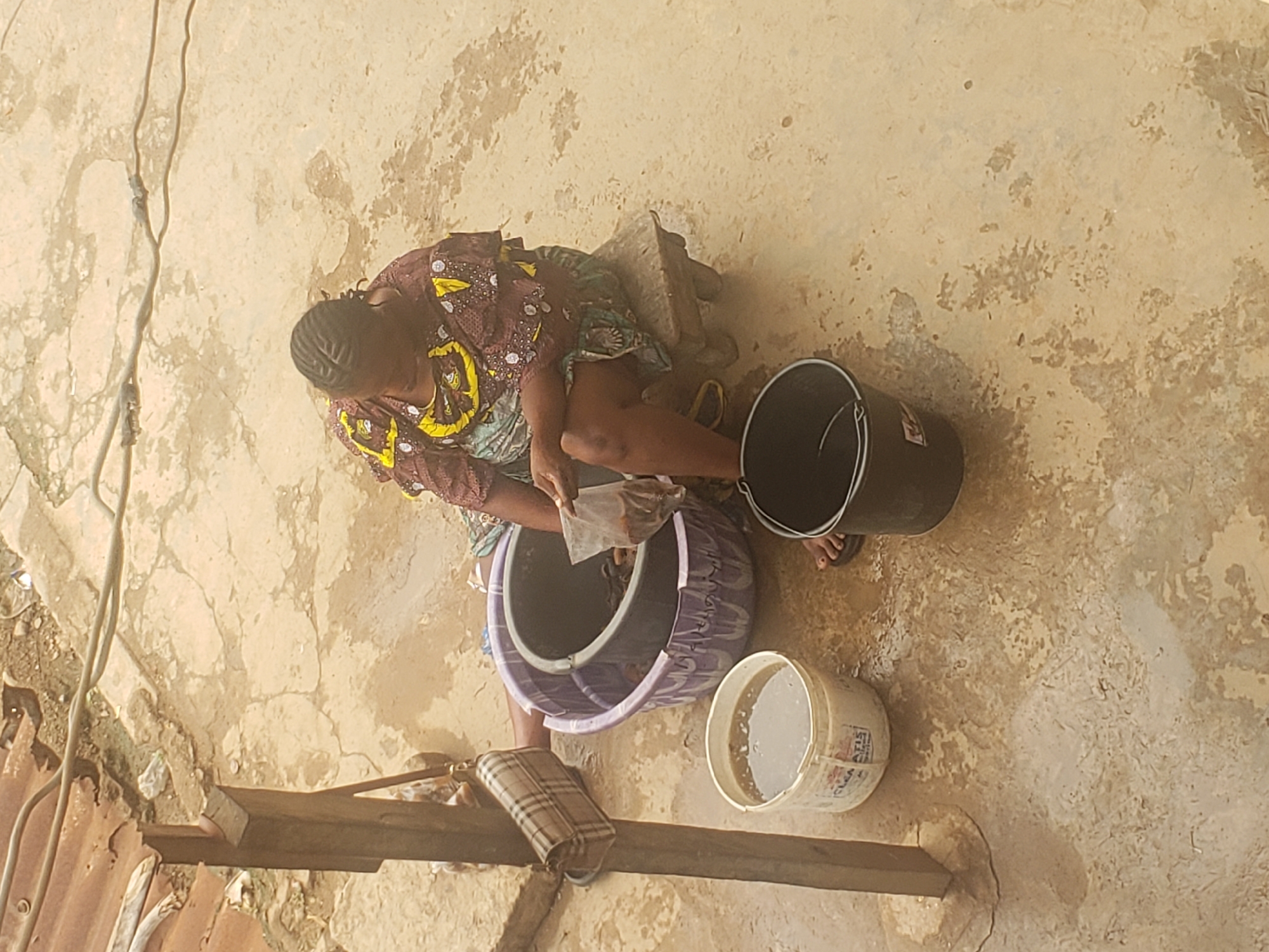 A woman in Alalubosa, Nigeria, is sitting outside on a stool. She is wearing a patterned dress and flip-flops. She appears to be washing something in a large purple bucket with a white interior. There is a clear plastic bag in her hand and items in the bucket. A black bucket and a white bucket are nearby. A purse rests on a piece of wood on the ground. The ground is a light tan color with cracks and stains. There are some wires visible.