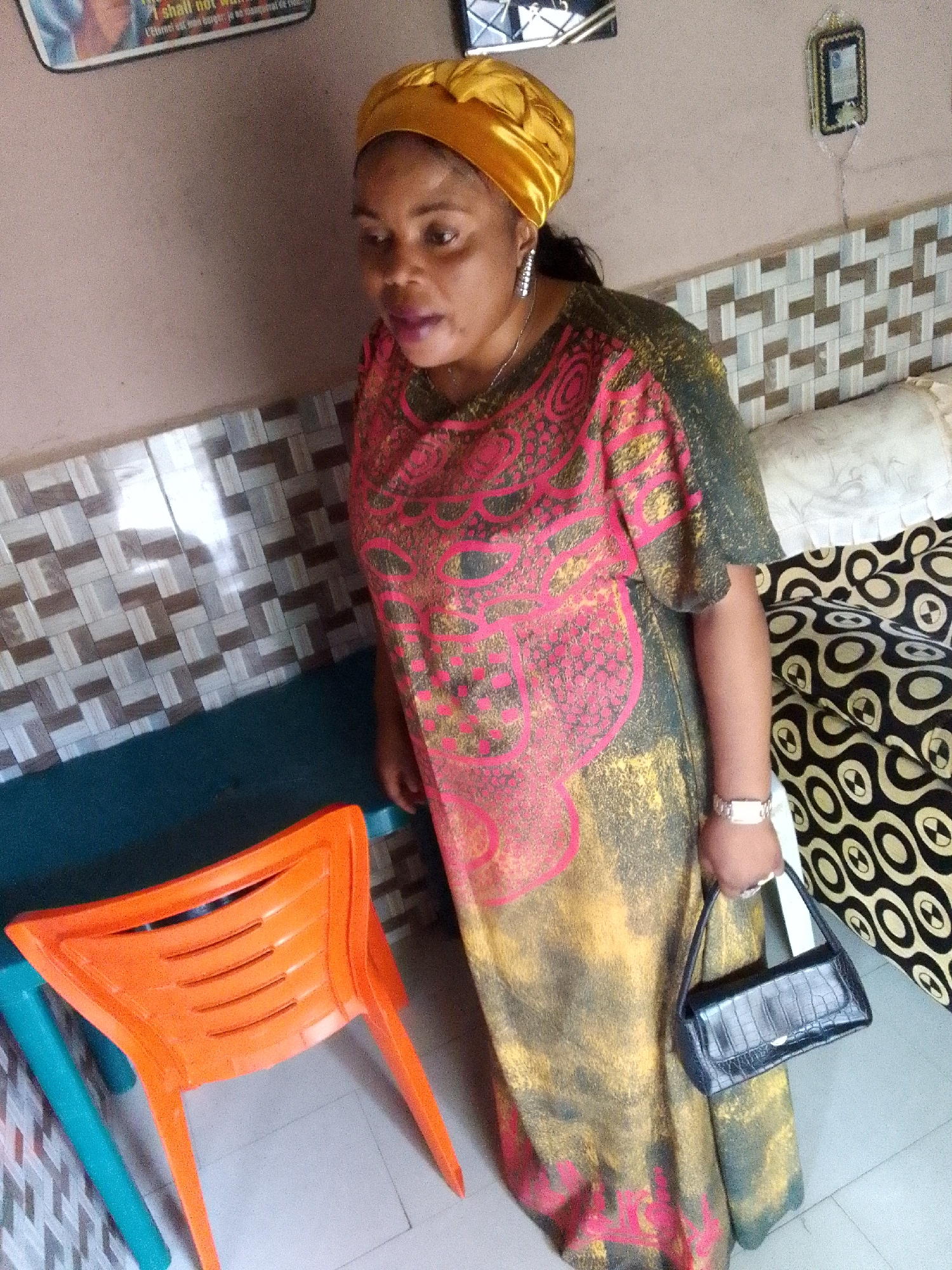 A woman wearing a yellow headscarf and a colorful dress stands next to an orange plastic chair. She is holding a black handbag. The background includes a wall with a patterned tile accent, a couch with a patterned cover, and a framed picture on the wall. The scene appears to be indoors, possibly in a residential setting in Jos, Nigeria.