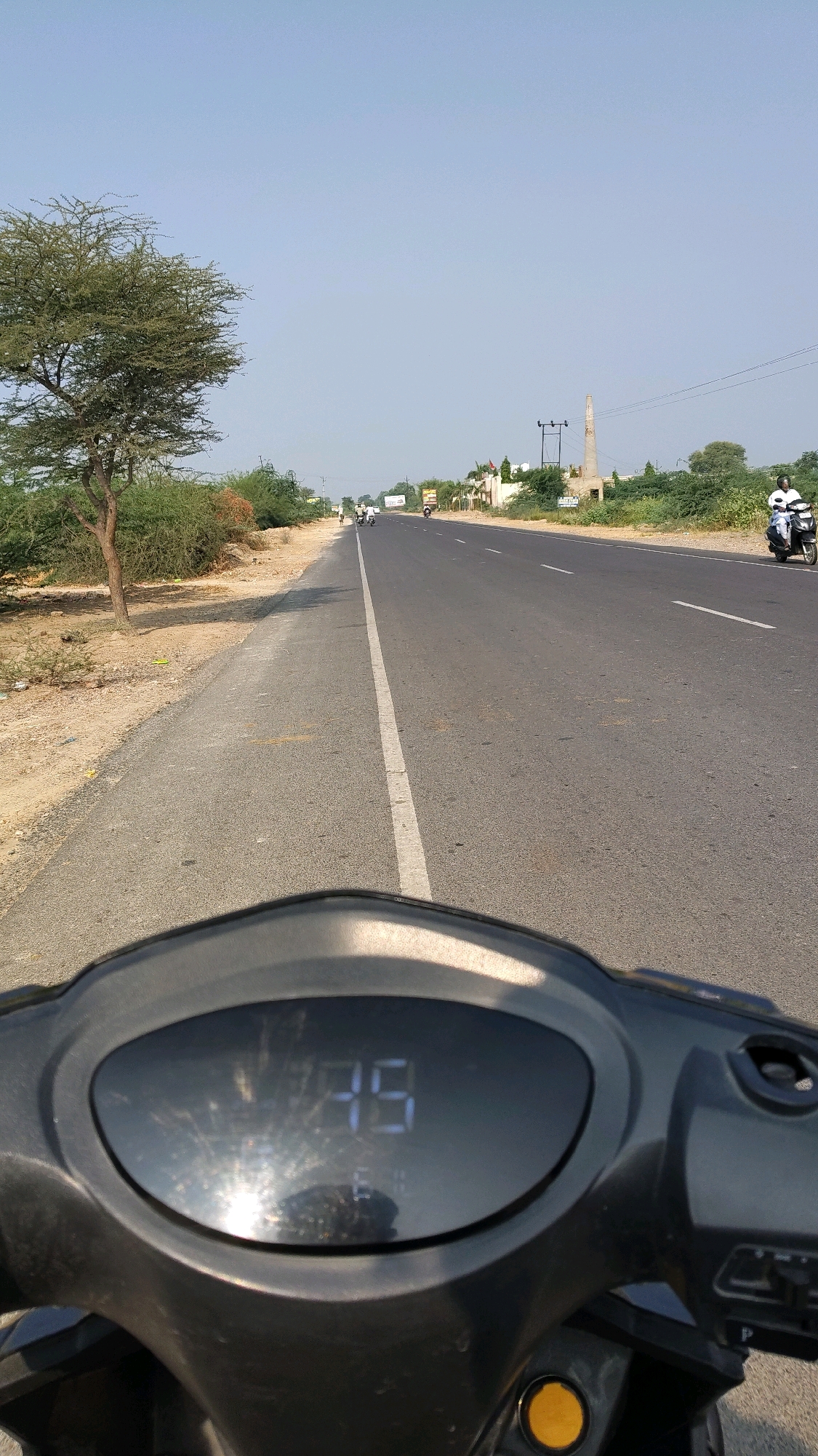 The image depicts a view from a scooter on a two-lane paved road in Sangawali, India. The road has a white line dividing the lanes and extends into the distance under a clear blue sky. On the left side of the road, there is a dirt shoulder with sparse vegetation and a large tree with sparse foliage. On the right side of the road, there are more trees and some buildings in the distance, including what appears to be a tall, slender, unfinished concrete structure, possibly a chimney or a monument, and an electricity pole with wires.
There are several vehicles on the road. In the foreground, the digital display of the scooter shows the number "75". Further down the road, there are a few motorcycles with riders, and on the far right, a person is riding a black and white scooter. The overall scene suggests a sunny day in a rural or semi-rural area.