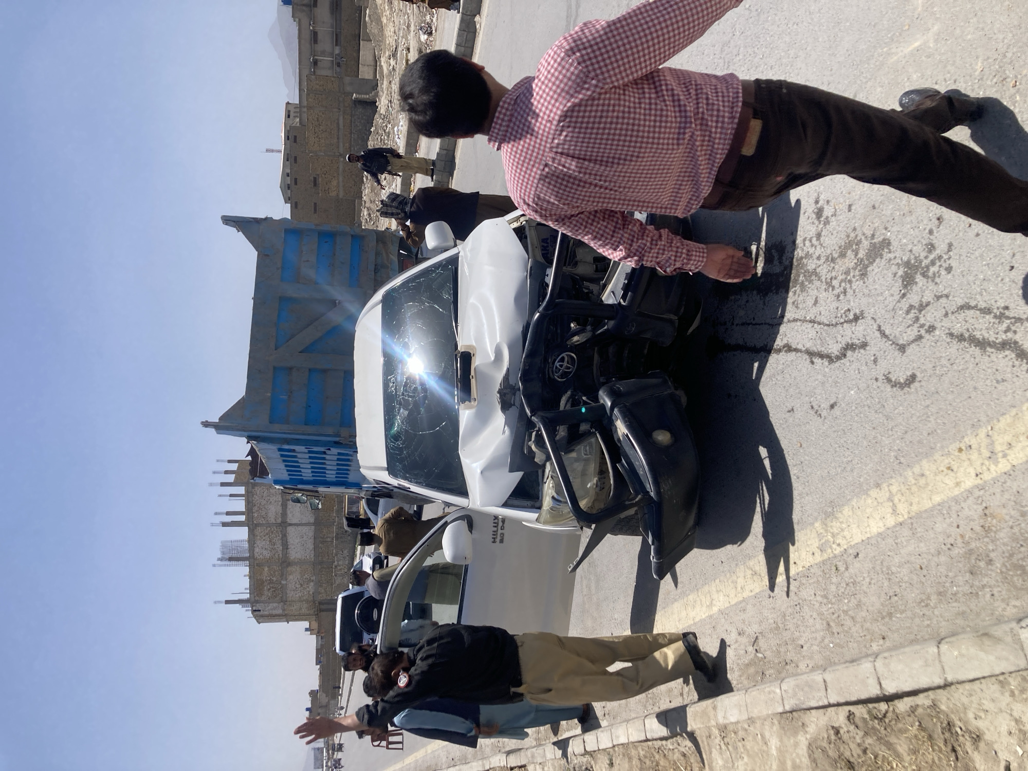 The image shows the aftermath of a vehicle collision in Quetta City Tehsil, Pakistan. A white Toyota Hilux pickup truck has sustained significant front-end damage, including a shattered windshield and a crumpled front bumper. The truck appears to have collided with a blue and white commercial vehicle, partially visible behind the Hilux.
Several individuals are present at the scene. One man wearing a red and white checkered shirt and brown pants is standing near the damaged truck. Several other people stand along the side of the road.
The surrounding environment shows buildings under construction. The sky is clear and the time of day appears to be daytime.