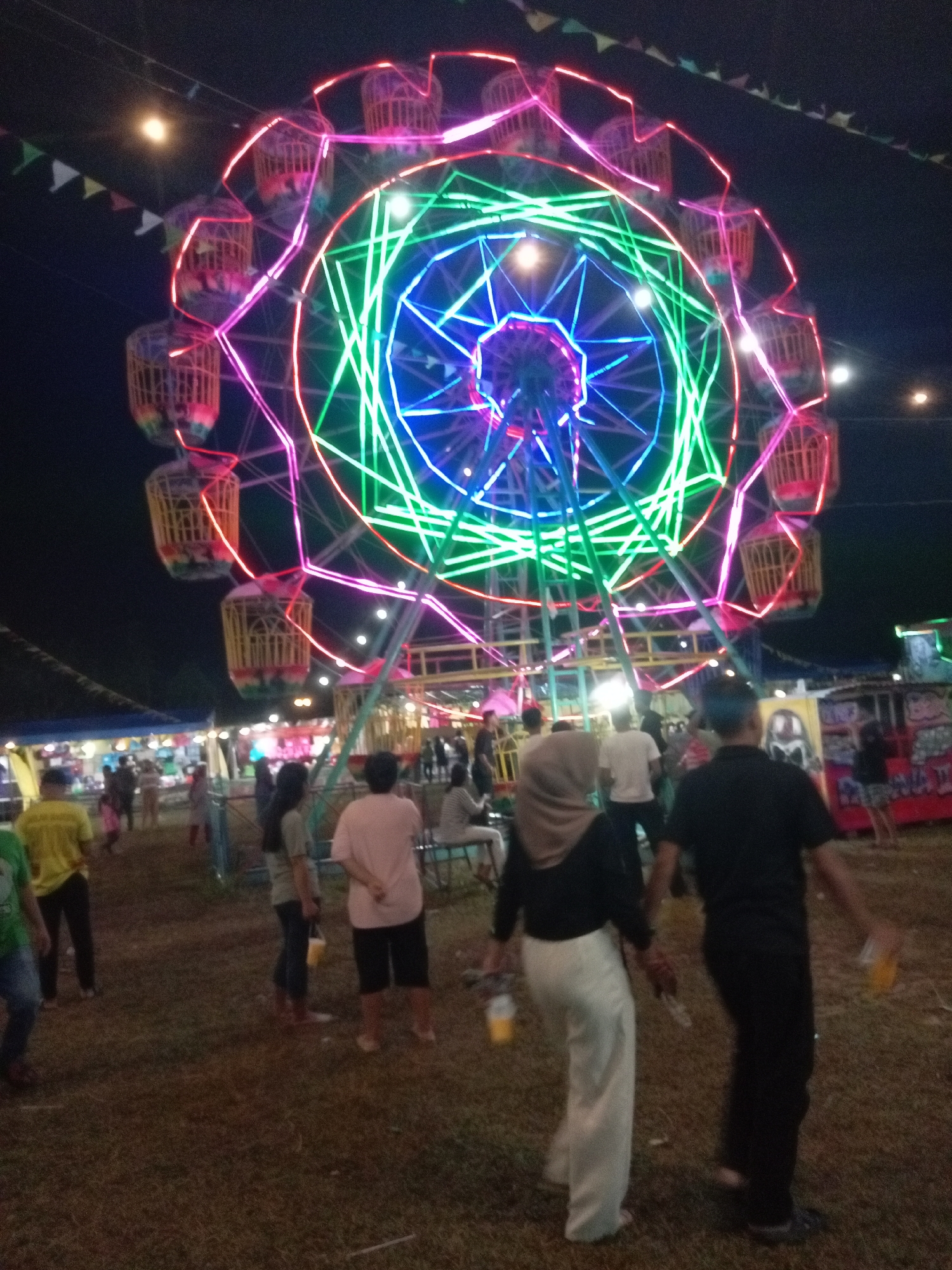 The image depicts a brightly lit Ferris wheel at night, likely at a carnival or fairground. The Ferris wheel is adorned with a vibrant display of neon lights in various colors, including pink, green, and blue, forming intricate geometric patterns. Several gondolas, appearing to be basket-like structures, are suspended from the wheel, also outlined with neon lights. In the background, there are tents and other fairground attractions. A group of people are visible in the foreground and mid-ground, milling around the area. Some individuals are walking, while others appear to be standing and observing. The ground is covered in dry grass, and the overall atmosphere suggests an outdoor evening event. The presence of a moon is also notable in the night sky. Without specific landmarks or textual clues, identifying the exact location in Batam, Indonesia is not possible from the image alone.