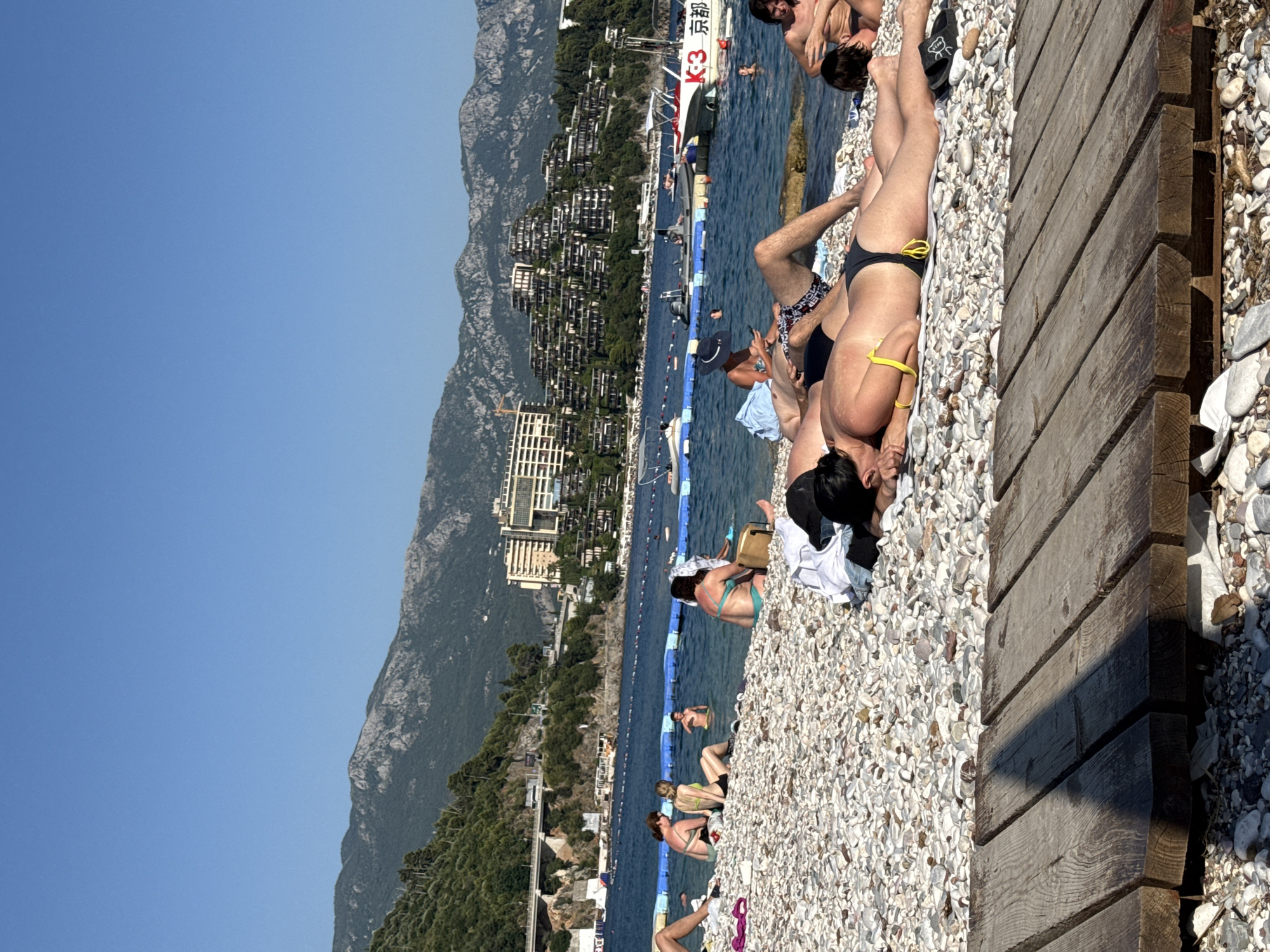 The image depicts a sunny day at a pebble beach in Budva, Montenegro. Several people are seen relaxing on the shore and in the water. In the foreground, a woman is sunbathing on a white towel, wearing a black bikini with yellow accents. To her left, another person is partially visible, also sunbathing. The beach is covered with smooth, light-colored pebbles. A wooden walkway runs along the right side of the frame.
In the background, the Adriatic Sea is visible, with a boat and a floating barrier marking a swimming area. Further back, a hillside is densely covered with trees and tiered buildings, characteristic of a coastal resort town. A large mountain range forms the backdrop, with a clear blue sky overhead. The scene suggests a leisurely vacation atmosphere, with people enjoying the warm weather and the coastal scenery. There are no discernible emotions visible in the individuals' poses, but the overall setting indicates a relaxed and pleasant environment. A boat in the water has "K3 京都" written on its side in red lettering.