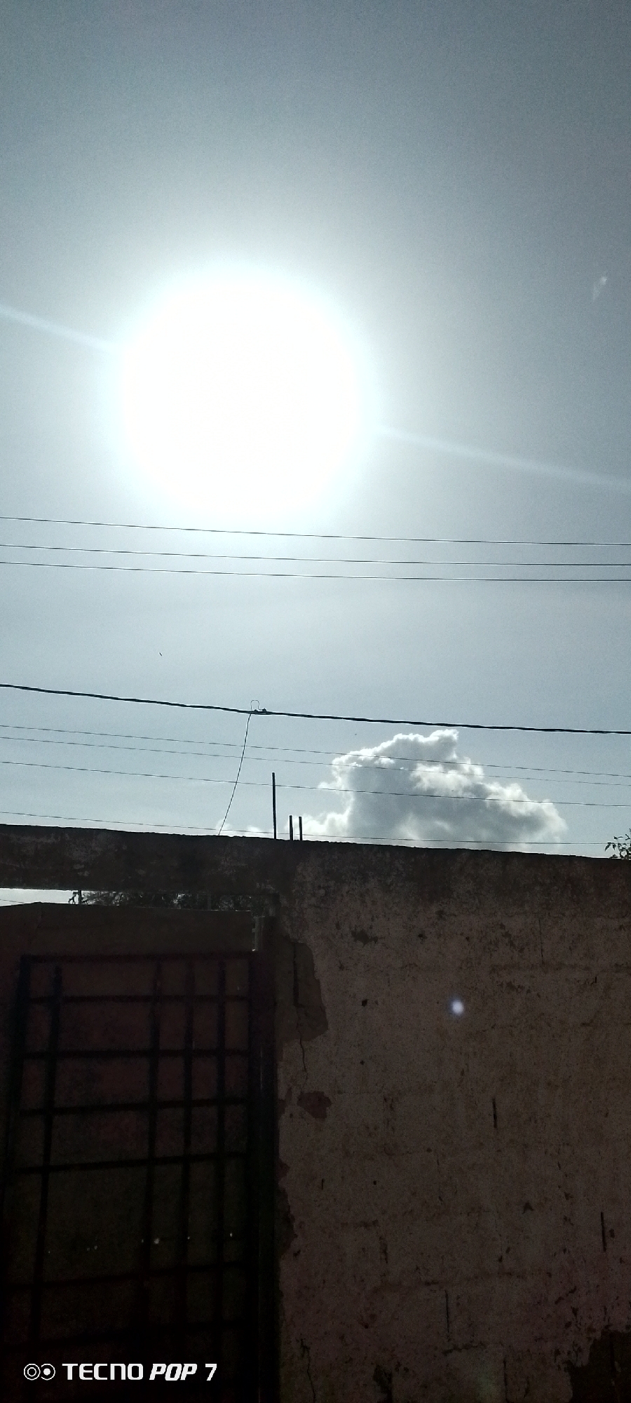 The image shows an exterior view in Altagracia, Venezuela. The sun is high in the sky, obscuring details. A weathered wall and barred window are in the foreground. Power lines stretch across the middle of the image, with a single cloud visible behind the wall. The lower portion of the image is in shadow. A "TECNO POP 7" logo is visible in the bottom left corner.