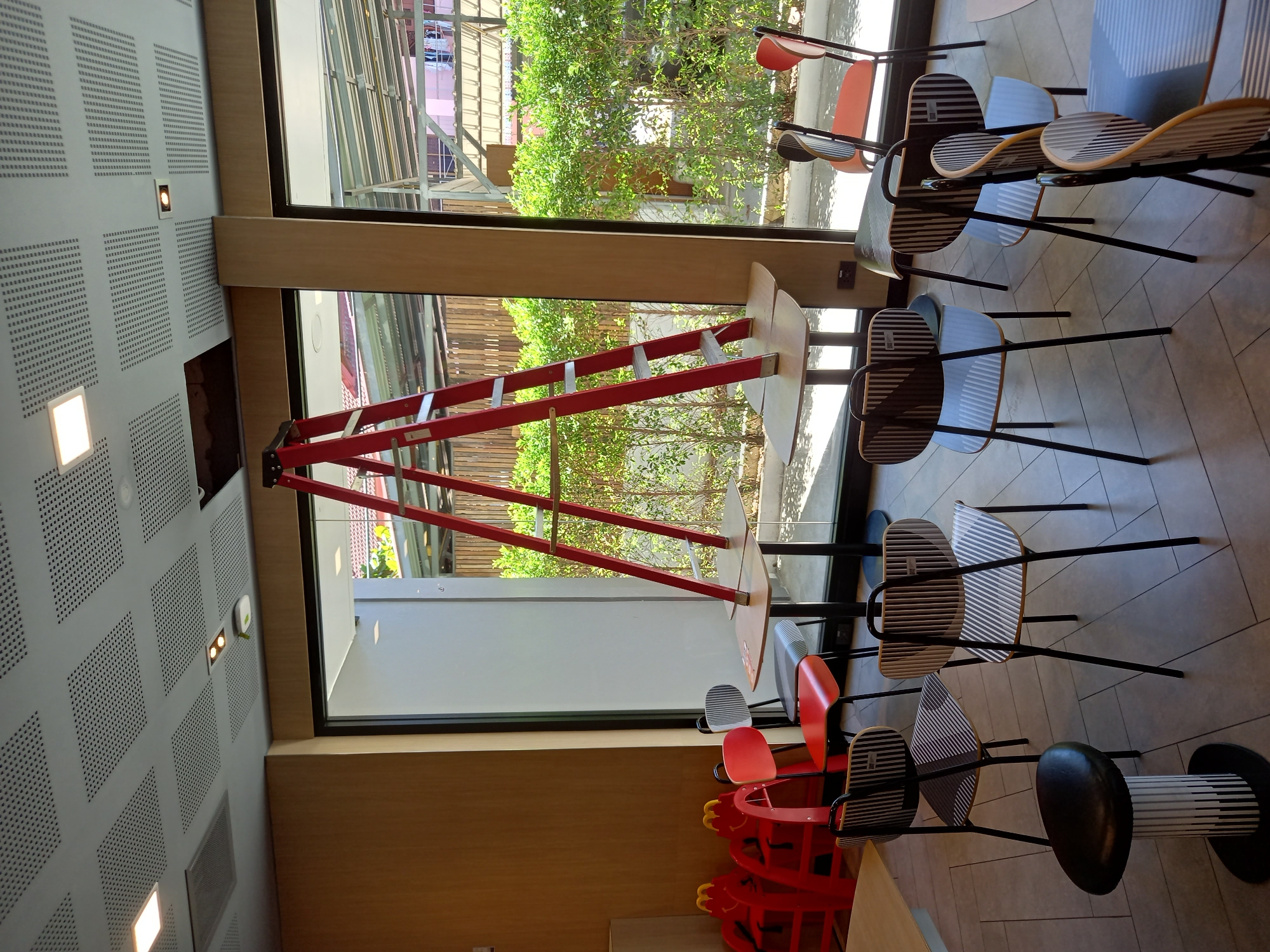 In Baan Bo Phut, Thailand, a McDonald's restaurant dining area is undergoing some form of maintenance or renovation. A bright red A-frame ladder is prominently displayed, straddling a light-colored table. The table itself appears to be two separate pieces, with the ladder legs supporting them. Surrounding the table are several chairs of various designs. Some are modern and sleek with striped backrests and seats, while others are a vibrant red. In the background, through large windows, lush green foliage and outdoor structures are visible, suggesting a bright, sunny day. To the left, there are children's high chairs or booster seats, also in red, with Mickey Mouse-like ears. The floor is tiled in a light gray pattern. The ceiling features square acoustic panels with perforations and embedded lighting fixtures. There are no people visible in the immediate scene, but the presence of the ladder and the arrangement of furniture suggest ongoing work within the establishment.