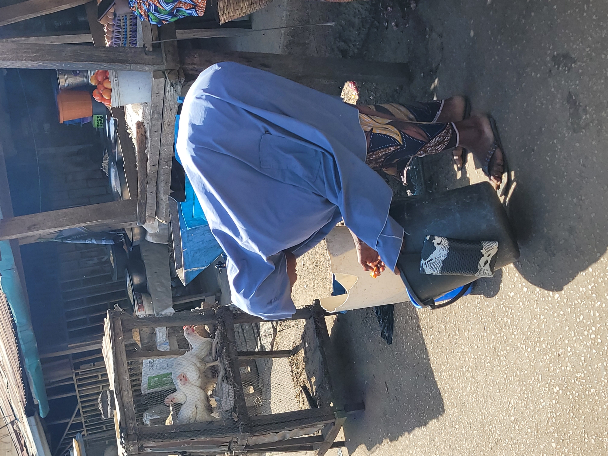 The image depicts an outdoor market scene, likely in Jos, Nigeria, during daylight hours, indicated by the strong shadows.
**Subjects:**
A person wearing a light blue, long-sleeved garment is central to the image, bent over with their back towards the viewer, holding a small orange item in their right hand. The lower legs and feet of another individual, clad in patterned trousers and dark flip-flops, are visible to the right. To the left, a rustic wooden cage holds three live white chickens. Surrounding the scene are various market items, including a white bucket filled with red tomatoes, an orange bucket, and a green bucket. A dark-colored, cylindrical container rests on the ground, with a small black and white patterned object placed on its rim. The market features makeshift wooden structures, serving as stalls or shelters.
**Activity or Event:**
The person in blue appears to be engaged in an activity involving the dark container, possibly picking up or placing an item, as their hand holds an orange object over it. The presence of goods like tomatoes and live chickens suggests active commerce in a market setting.
**Notable Details:**
Bright sunlight casts distinct shadows on the ground and market structures, indicating it is likely midday or late afternoon. The ground is a textured, unpaved surface typical of open-air markets. The overall setup, including the wooden stalls, types of goods, and clothing, aligns with an informal market environment common in regions like Jos, Nigeria.
**Visible Text:**
Inside the chicken cage, a white sack shows partially legible text that appears to be "CHICA" followed by indistinct letters, possibly "GRO."