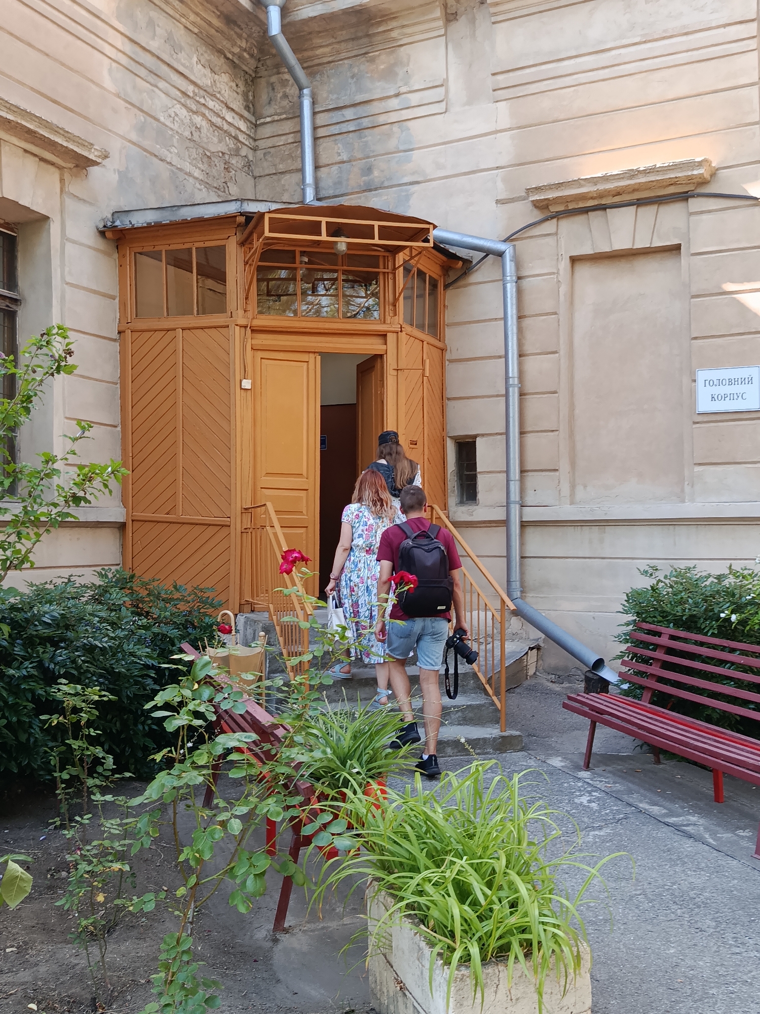 The image shows the exterior of a building with a wooden porch and entrance. Three individuals are walking up the stairs towards the entrance. The first person, on the left, is wearing a floral dress and carrying a white bag. The second person, in the middle, is wearing a dark backpack and carrying a camera. The third person, on the right, is wearing denim shorts and a maroon t-shirt, also carrying a camera. To the right of the stairs, there is a red bench. Green bushes and plants are visible in the foreground and to the left of the entrance. A sign on the wall to the right reads "ГОЛОВНИЙ КОРПУС". The building appears to be old, with textured walls and a grey downspout. The lighting suggests it is daytime.