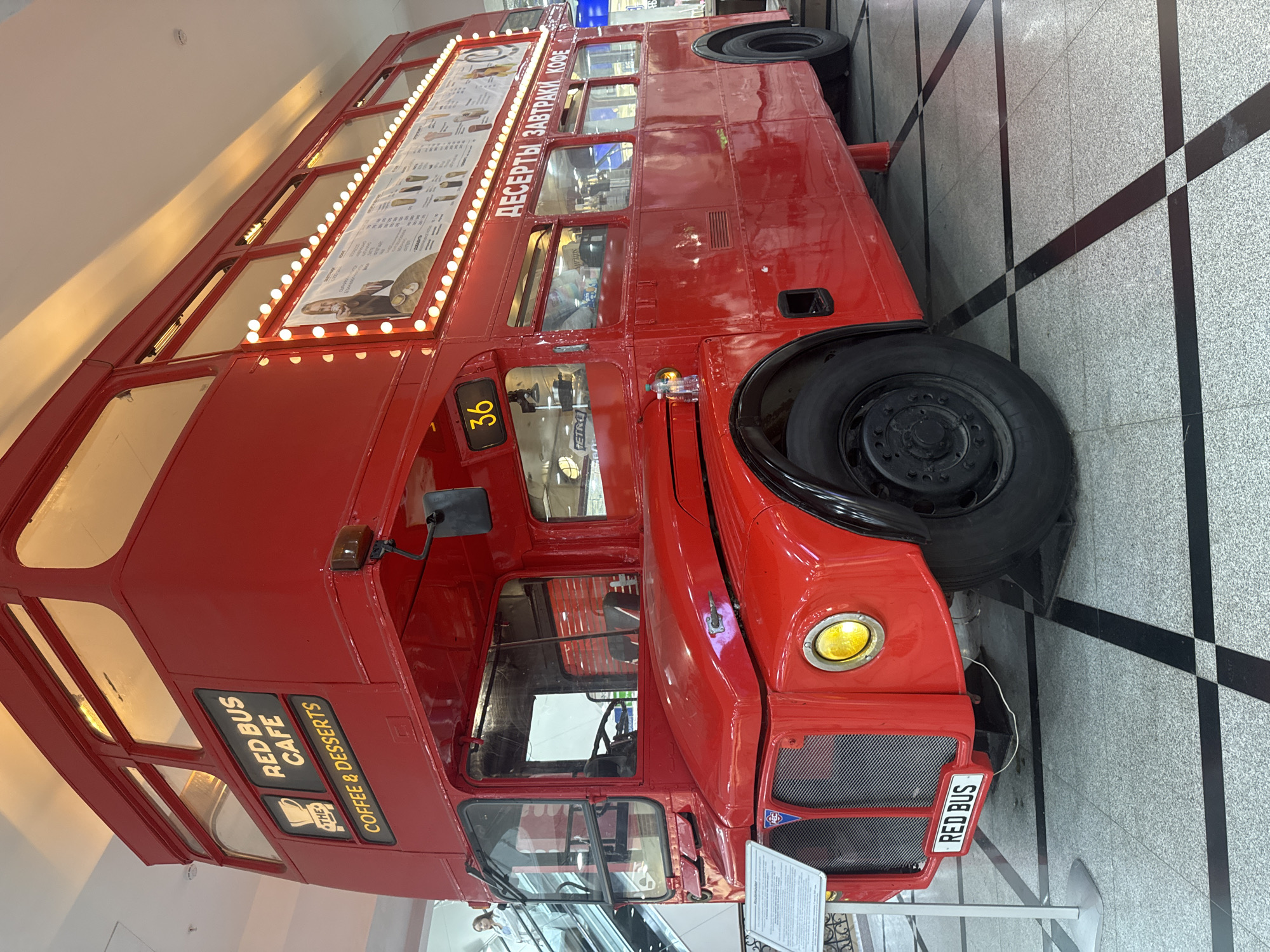 The image shows a red double-decker bus, repurposed as "RED BUS CAFE," with "COFFEE & DESSERTS" written on its side. The bus is indoors, on a patterned tiled floor with black lines. It is illuminated by warm interior lights and a string of lights along the upper level. The front of the bus features a license plate that reads "RED BUS" and a yellow headlight. A menu board is visible on the upper level, displaying Cyrillic text that translates to "DESSERTS, BREAKFAST, COFFEE." A woman in a light-colored jacket is seen on an escalator in the background, to the left of the bus. The overall scene appears to be a commercial space, possibly a shopping mall or a public venue in Yekaterinburg, Russia.
