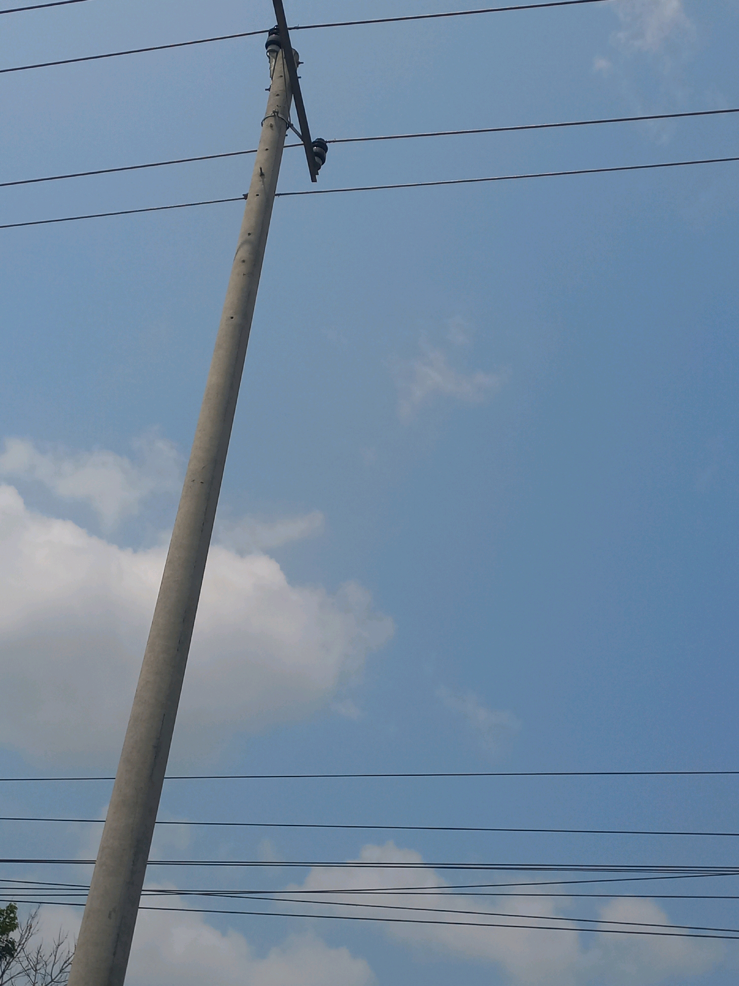 The image captures a concrete utility pole leaning against a clear blue sky with scattered white clouds. Several dark electrical wires stretch horizontally across the frame, appearing to be supported by the pole. The pole itself is textured and appears to be made of reinforced concrete, with visible insulators attached near its top. The image does not contain any people, animals, or any specific identifying features that would indicate its location in Baturetno, Indonesia. The focus is on the utility pole and the overhead wires against the backdrop of the sky and clouds.
