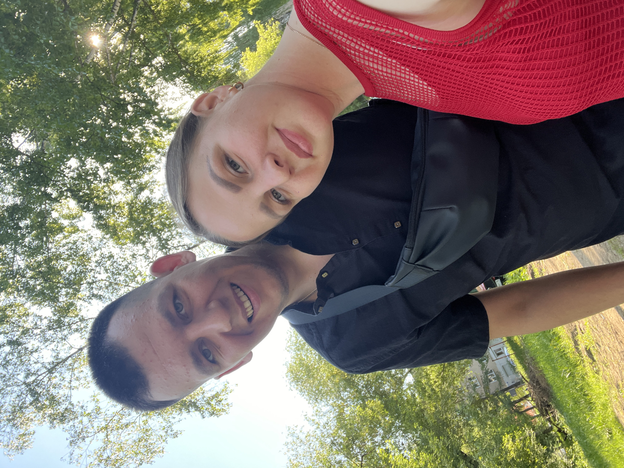 A selfie taken outdoors in Izhevsk, Russia, depicts a man and a woman. The woman is wearing a red, mesh top and a black jacket with a strap. The man is wearing a black shirt. They are standing close together and smiling. Trees are visible in the background, along with grass and a building. The sun is shining through the trees.