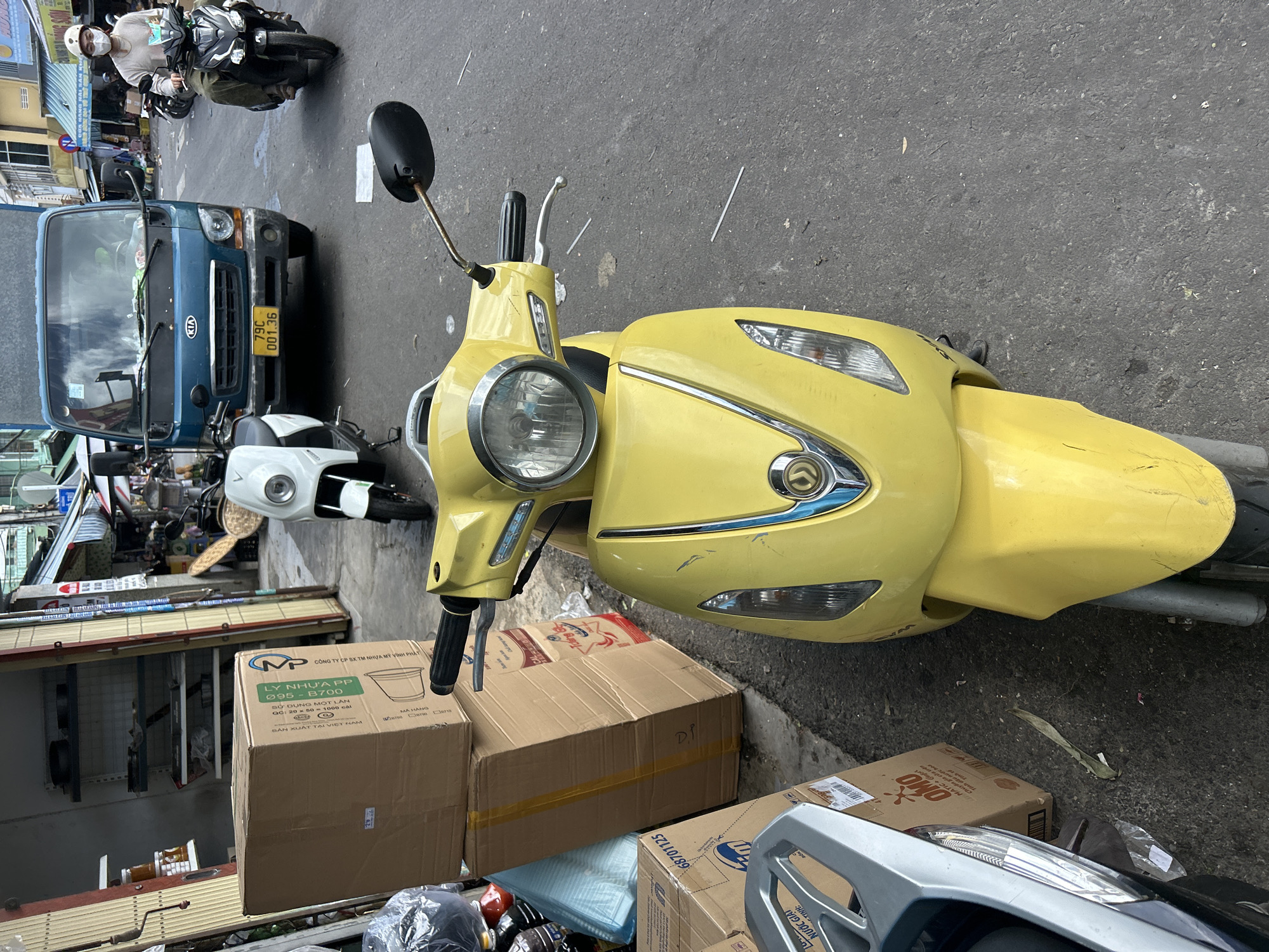 This daytime image captures a busy street scene in Nha Trang Ward, Vietnam, featuring various vehicles and stacked commercial goods. In the foreground, a vibrant yellow scooter is prominently parked, its headlight and handlebars visible. To its left, a blue Kia truck is stationary, displaying a Vietnamese license plate "79C 001.36". A white scooter is parked behind the truck. In the background, a person wearing a helmet and face mask drives a black scooter down the street. Another silver scooter is partially visible in the bottom right.
Several cardboard boxes are placed around the yellow scooter, indicating either temporary storage or goods being transported. Visible text on these boxes includes Vietnamese phrases such as "CÔNG TY CP SX TM NHỰA MỸ VĨNH PHÁT", "LY NHỰA PP 095-B700" (likely referring to polypropylene plastic cups), "SỬ DỤNG MỘT LẦN" (single-use), and "SẢN XUẤT TẠI VIỆT NAM" (Made in Vietnam). Additionally, packaging for "OMÔ NƯỚC GIẶT" (Omo laundry detergent) is discernible. The street appears to be asphalt, and glimpses of buildings and commercial structures are visible in the background, typical of an urban setting. The clear lighting suggests it is a bright, sunny day.