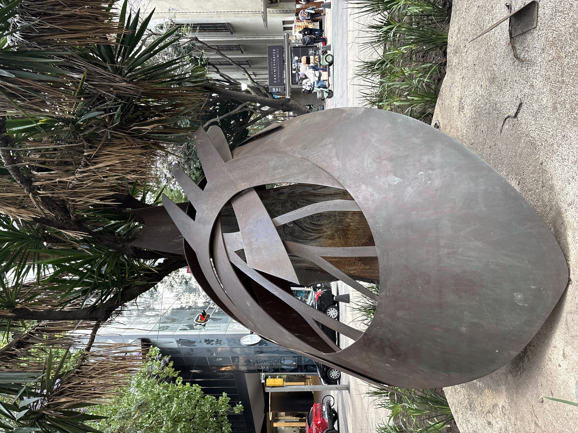 Here is a detailed description of the image:
**Scene:** A ground-level, daylight outdoor shot showcasing a large metal sculpture. The sculpture is abstract, featuring curved and angular metal pieces arranged within a circular frame. The metal appears weathered or patinated, with a dark, matte finish.
**Location:** The scene is located in Mexico City, Mexico. This is indicated by the signage "KARIMIDERDINDER, LA VERDADERA MATRIOSHKA" in the background.
**Environment:** The sculpture is situated on a concrete platform within a green, urban environment. A tall yucca-like tree or shrub is positioned to the left of the sculpture. Various other plants and low shrubs surround the sculpture and platform. Buildings are visible in the background, including shops with glass storefronts. Street activity can be observed, with people walking and a golf cart driving by.
**Objects:** Apart from the sculpture, the scene includes buildings, trees, shrubs, street signs, sidewalks, and vehicles.
**Overall Impression:** The image presents a modern sculpture integrated into the urban landscape of Mexico City. The juxtaposition of the abstract art, natural greenery, and bustling city atmosphere creates a dynamic and visually interesting composition.