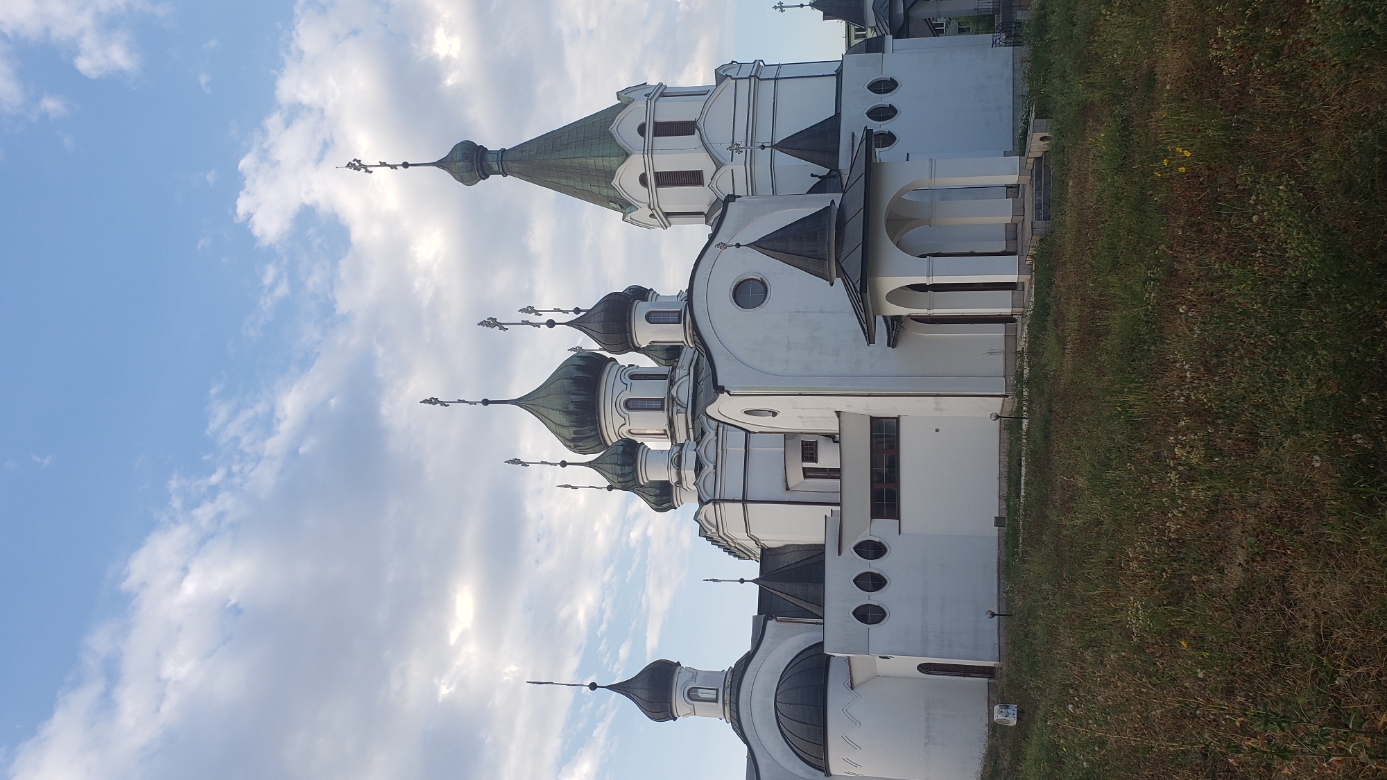 This is a vertical, full shot of the Orthodox Church of the Holy Spirit in Prešov, Slovakia, under a partly cloudy sky. The white church has multiple green domes topped with crosses. The main structure is large and white with a prominent bell tower on the right. A smaller, domed section is visible on the left, and an arched portico is in the foreground. The church is surrounded by green grass and some dry weeds. The lighting suggests it is either early morning or late afternoon. There are no people visible in the image.