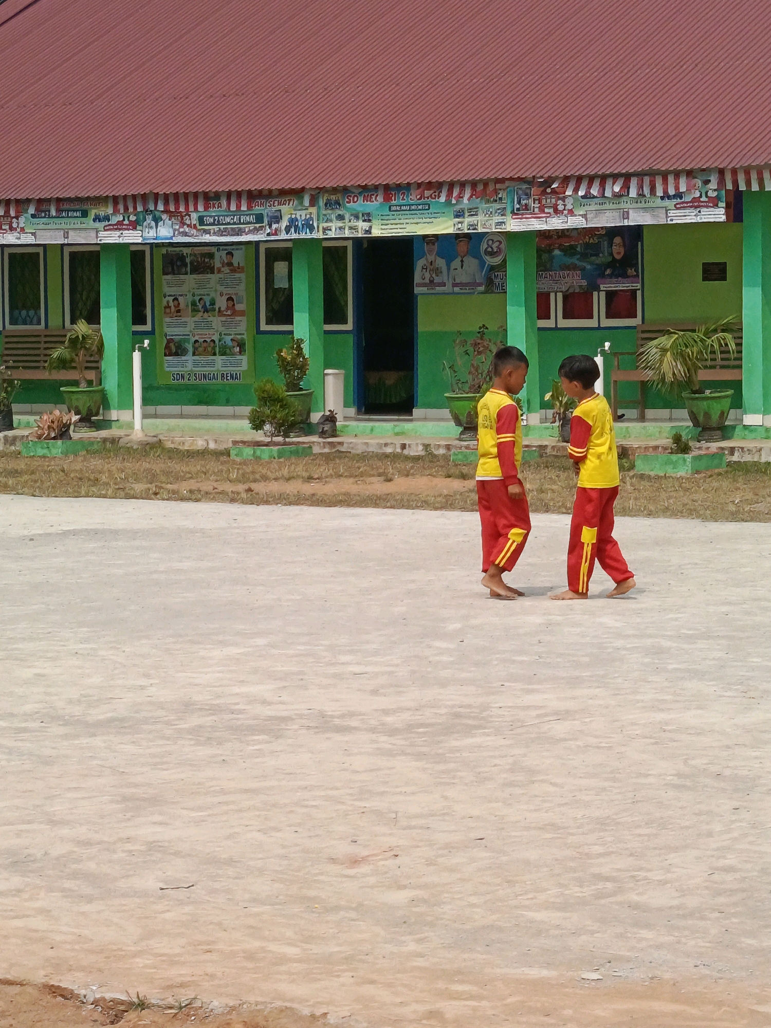 Two young boys, barefoot and dressed in matching yellow shirts with red collars and cuffs and red pants with yellow stripes, stand facing each other on a light gray paved surface. The boy on the left is looking down at his feet, while the boy on the right is looking at the boy on the left. Behind them, a vibrant green building with a red, corrugated metal roof is visible. The building has several windows and doors, and a banner runs along the top of the facade. Posters are displayed on the walls, and potted plants are arranged on the ground in front of the building. The environment appears to be an outdoor schoolyard in Megang Sakti IV, Indonesia, characterized by the building's architecture and the presence of a school banner. The ground is a mix of paved areas and dry, patchy grass.