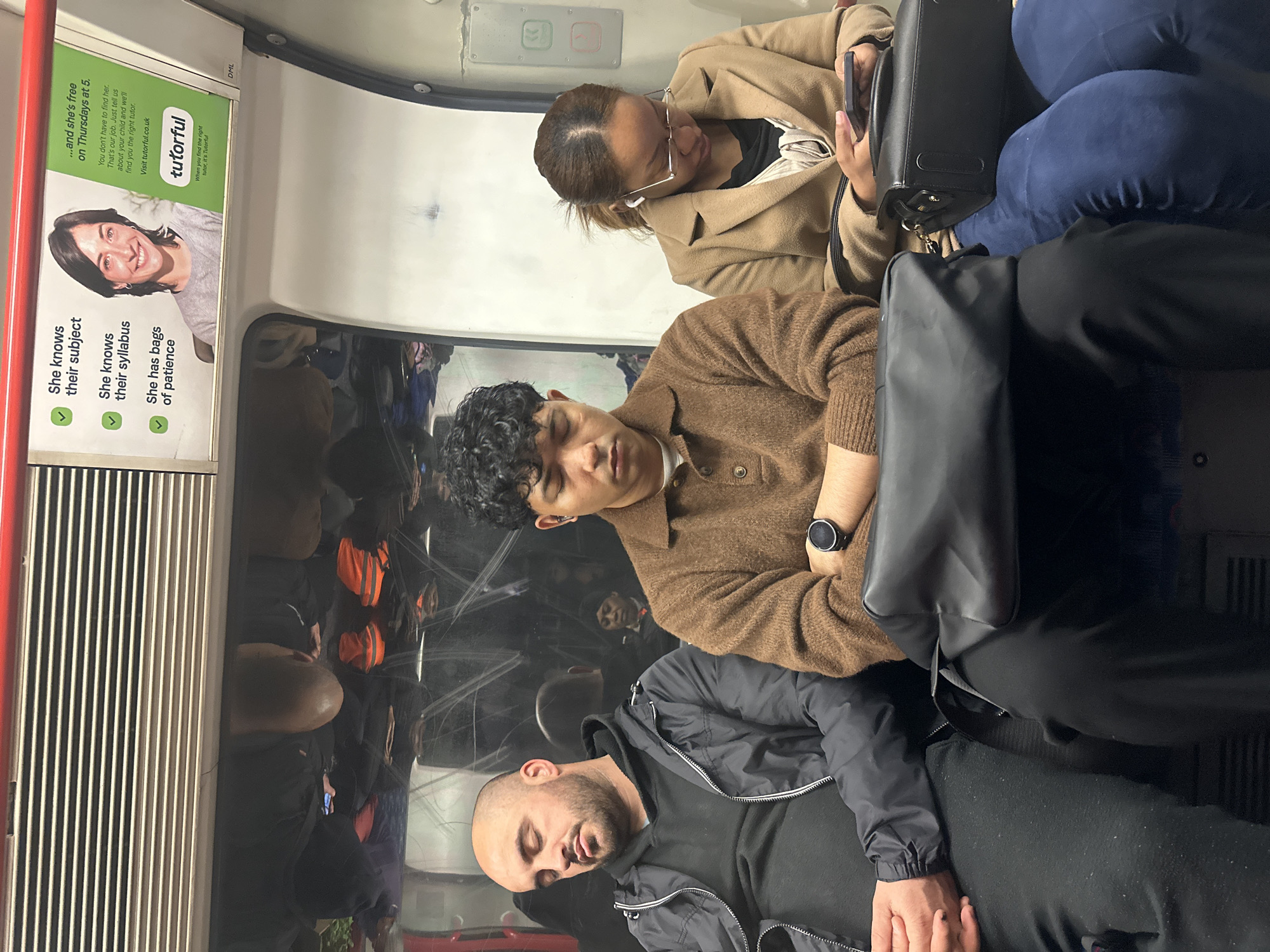 An interior shot aboard a London Underground train captures several passengers during their commute in the United Kingdom. The image, taken from a slightly elevated perspective, shows the train carriage with its characteristic advertising panels and reflective surfaces.
In the foreground, a man with a shaved head and beard, wearing a black jacket over a dark top, is visibly asleep with his eyes closed and mouth slightly ajar. His hands are clasped over a black backpack resting on his lap. Next to him, positioned slightly further back, is another young man with dark, curly hair, dressed in a brown knitted sweater. He is also sleeping with his eyes shut and wears a black wristwatch on his left wrist. A black handbag sits between these two men.
Further into the background, a woman with brown hair, wearing glasses and a light brown coat, is engaged with her smartphone, looking down at the device while wearing white earbuds. Beside her, another passenger wearing dark blue trousers is partially visible. The train window or reflective surface opposite these passengers shows clear reflections of numerous other commuters, some in orange high-visibility clothing, indicating a crowded environment.
A prominent green digital advertisement for 'tutorful' is displayed on the train wall. The ad features a smiling woman and promotional text that includes: 'She knows their subject,' 'She knows their syllabus,' 'She has bags of patience,' and '...and she's free on Thursdays at 5.' It further states: 'You don't have to find her. That's our job. Just tell us about your child and we'll find you the right tutor. Visit tutorful.co.uk.' Below the logo, it reads, 'When you find the right tutor, it's Tutorful.' A small 'DML' tag is also seen above the advertisement panel.
The scene conveys the common urban commuting experience, characterized by passengers resting or occupying themselves on a busy public transport service, likely during daytime given the bright interior lighting. The overall atmosphere suggests the routine and often quiet exhaustion of daily travel.