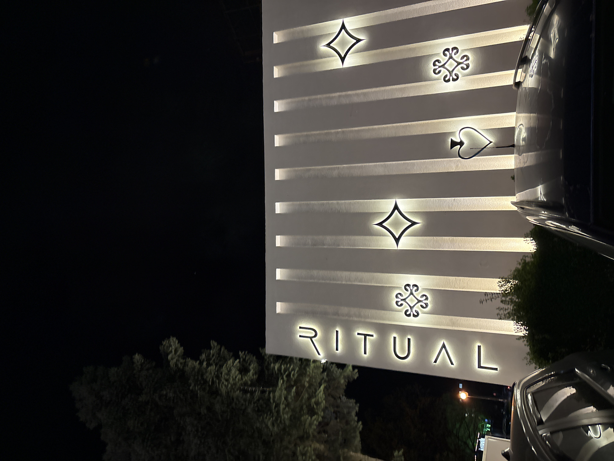 The image shows the exterior of a building at night, identifiable by the illuminated word "RITUAL" and card suit symbols on its facade. The building is light-colored, and the facade has horizontal grooves illuminated with light. A car is parked to the right, partially obstructing the view of the building. In the foreground, there are trees and bushes. The background is a dark night sky. The image is likely from Sofia, Bulgaria due to the distinctive "RITUAL" sign associated with that city.