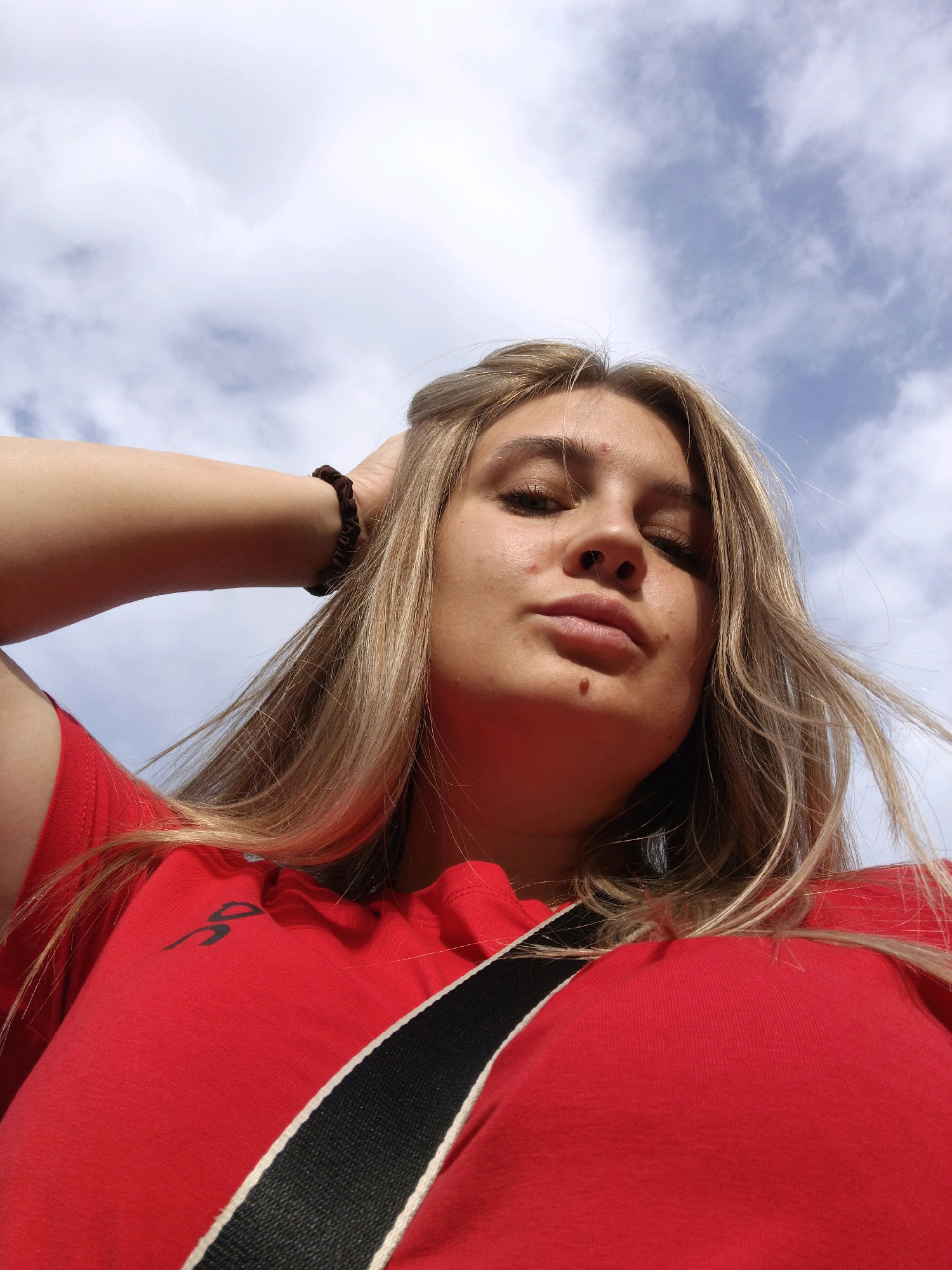 A low-angle, outdoor, waist-up shot features a young woman with blonde hair and a red shirt. She is looking down at the camera, with her right arm raised and resting behind her head. A dark scrunchie is visible on her wrist. A black and white strap is visible across her chest. The background is a bright blue sky with scattered white clouds. The image was taken on a sunny day. The subject appears to be in a relaxed and casual pose. There are no other individuals or identifiable structures visible in the image. The specific location in Zlatoust, Russia is not discernible from the visual information presented.