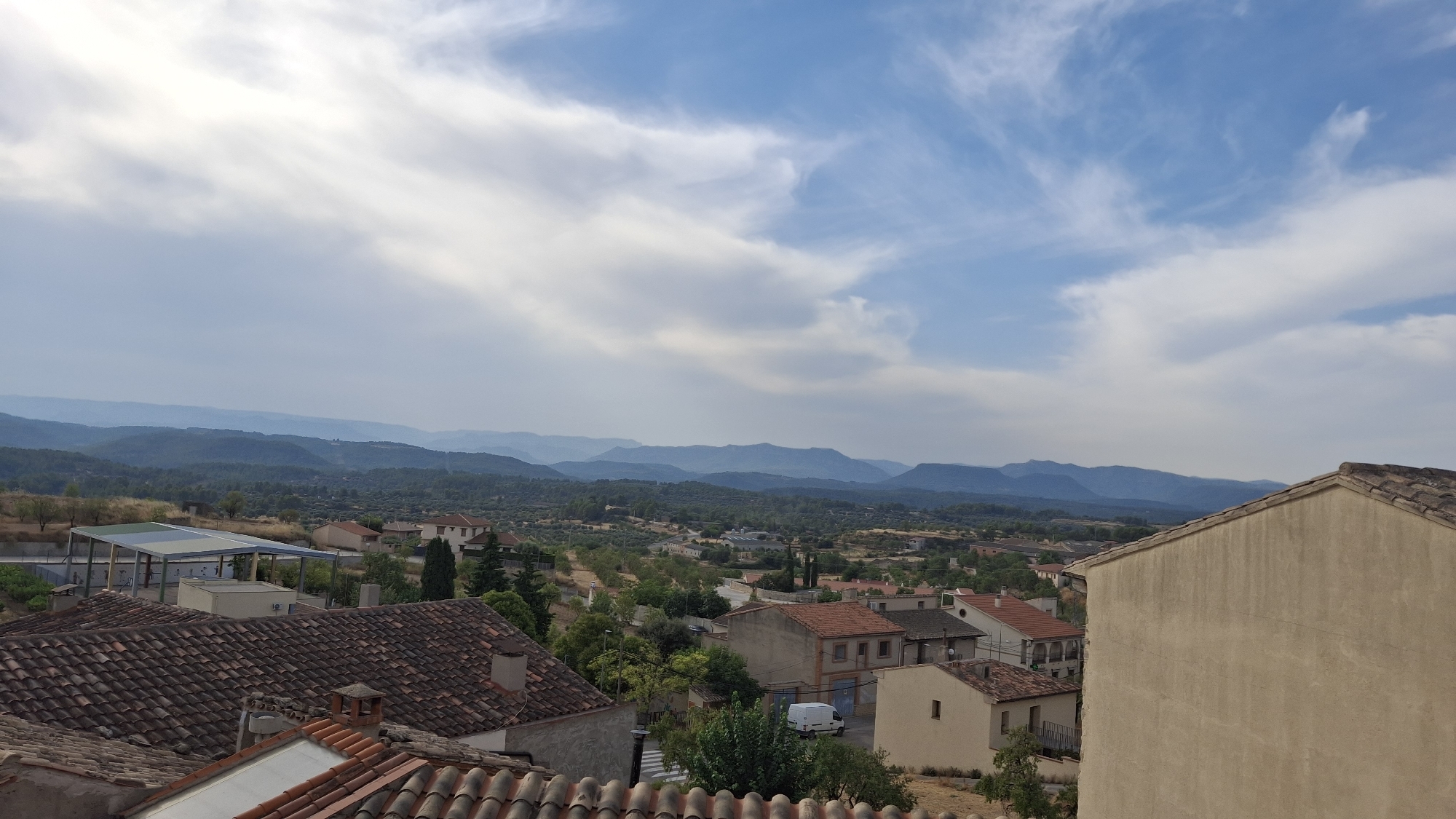 The image captures an elevated, panoramic view of the town of La Fresneda, Spain. In the foreground, terracotta tiled roofs of residential buildings are prominent, with the side of a light-colored building dominating the right edge. Beyond these immediate structures, the town unfolds with a mix of residential buildings featuring red-tiled roofs and stucco exteriors. A covered structure, possibly a sports facility with a green and blue roof, is visible in the mid-ground. The town is nestled within a valley, with rolling hills covered in dark green foliage and scattered trees extending towards the horizon. A distant mountain range, shrouded in a bluish haze, forms the backdrop under a sky characterized by scattered white clouds against a pale blue expanse. The overall scene depicts a tranquil, rural town surrounded by natural landscape under a bright sky.