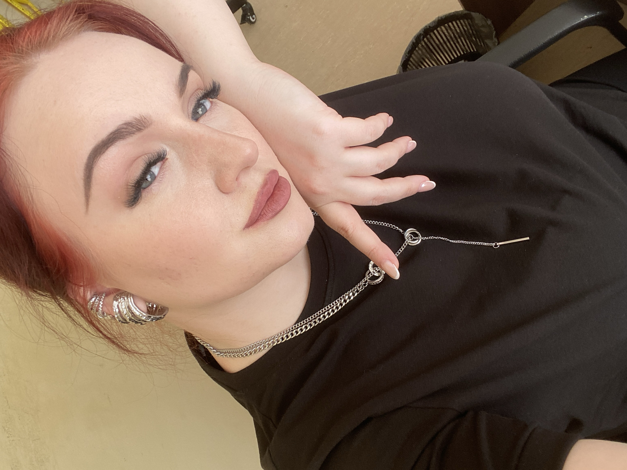 A woman with red hair and light skin poses for a selfie-style photo. She is wearing a black shirt, a silver chain necklace with ring pendants, and multiple silver earrings. Her left hand rests on her face. Her fingernails are manicured with a French tip. Her makeup includes eyeshadow, mascara, and lipstick.
It is not possible to determine the specific location in Chaikovsky, Russia, or any other city, from the information given in this image.