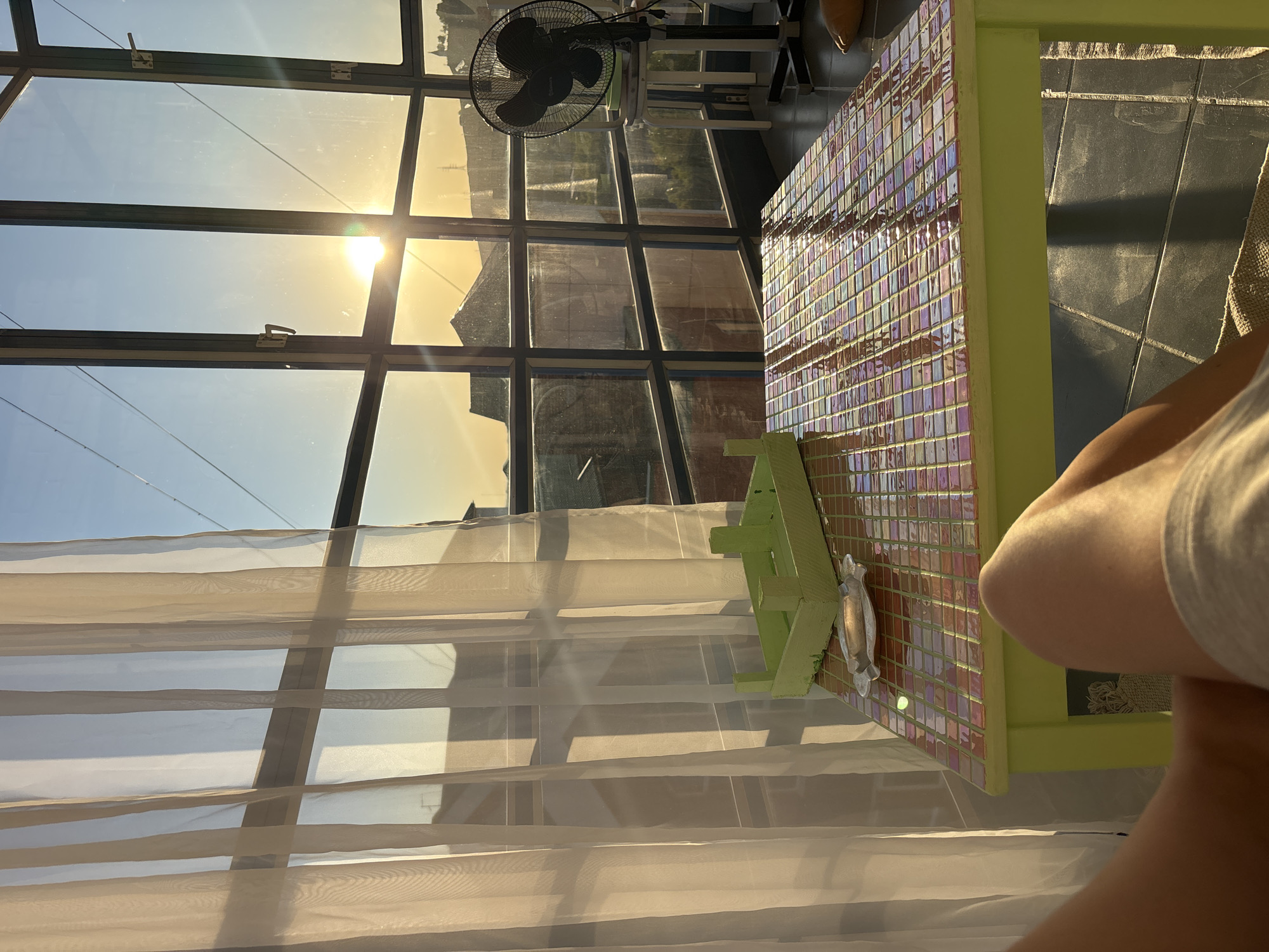 The image captures an indoor space, likely a sunroom or enclosed balcony in Krasnoyarsk, Russia, bathed in the warm light of a setting or late afternoon sun. A large, grid-framed window system dominates the left and central parts of the scene. The bright sun is visible through the glass, creating a strong glare and casting a golden hue across the room. The clear sky beyond the window appears light blue, transitioning to a golden yellow near the horizon.
An electric fan, black and with a visible guard, is either mounted or placed near the top of the window frame on the right side, partially silhouetted against the bright sky. Sheer, light-colored curtains hang over the lower section of the windows, diffusing the strong sunlight and creating soft folds.
To the right, a table with a vibrant, iridescent tiled surface and a light green frame occupies a significant portion of the foreground. Reflections shimmer on the multicolored tiles. On the table, there is a small, light green wooden stand and a metallic decorative object resembling a fish or stylized animal. The floor, visible in the lower right, consists of dark gray or black tiles. Part of a person's limb, likely a leg or arm, is visible in the bottom right corner, suggesting someone is seated or reclining, enjoying the peaceful and warm atmosphere of the sunny late afternoon. No discernible text is visible in the image.
