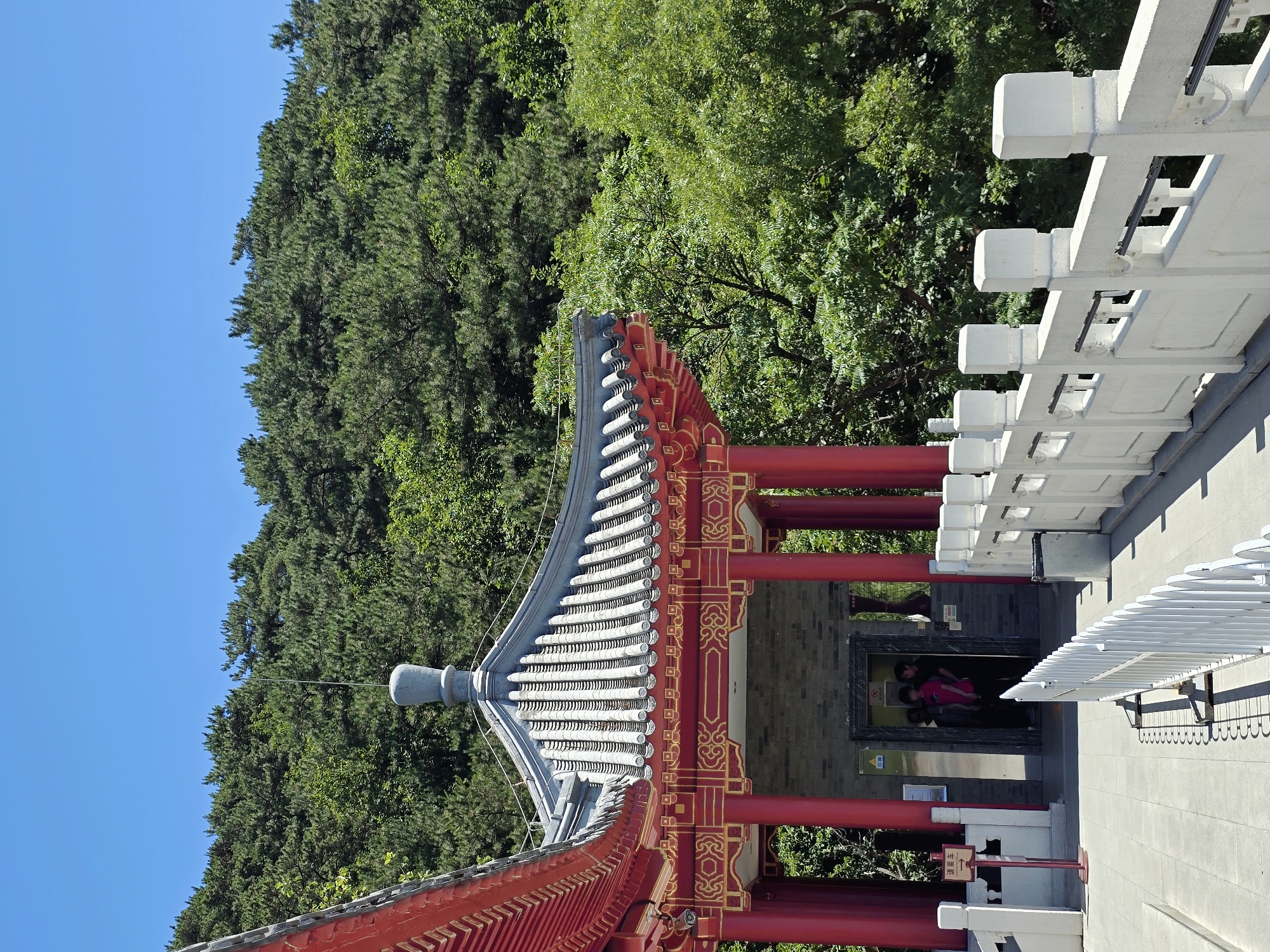 The image shows a traditional Chinese pavilion with red pillars and a gray tiled roof, set against a backdrop of lush green trees and a clear blue sky. The pavilion features ornate gold detailing and a prominent spire on its roof. In front of the pavilion, there is a paved walkway with a white railing. A few people are visible near an entrance to the pavilion, possibly an elevator, as they appear to be waiting. A sign with Chinese characters is mounted on a red post to the left of the walkway. The scene suggests a tourist destination in a natural setting, likely in Huairou District, China, given the architectural style and surrounding vegetation.