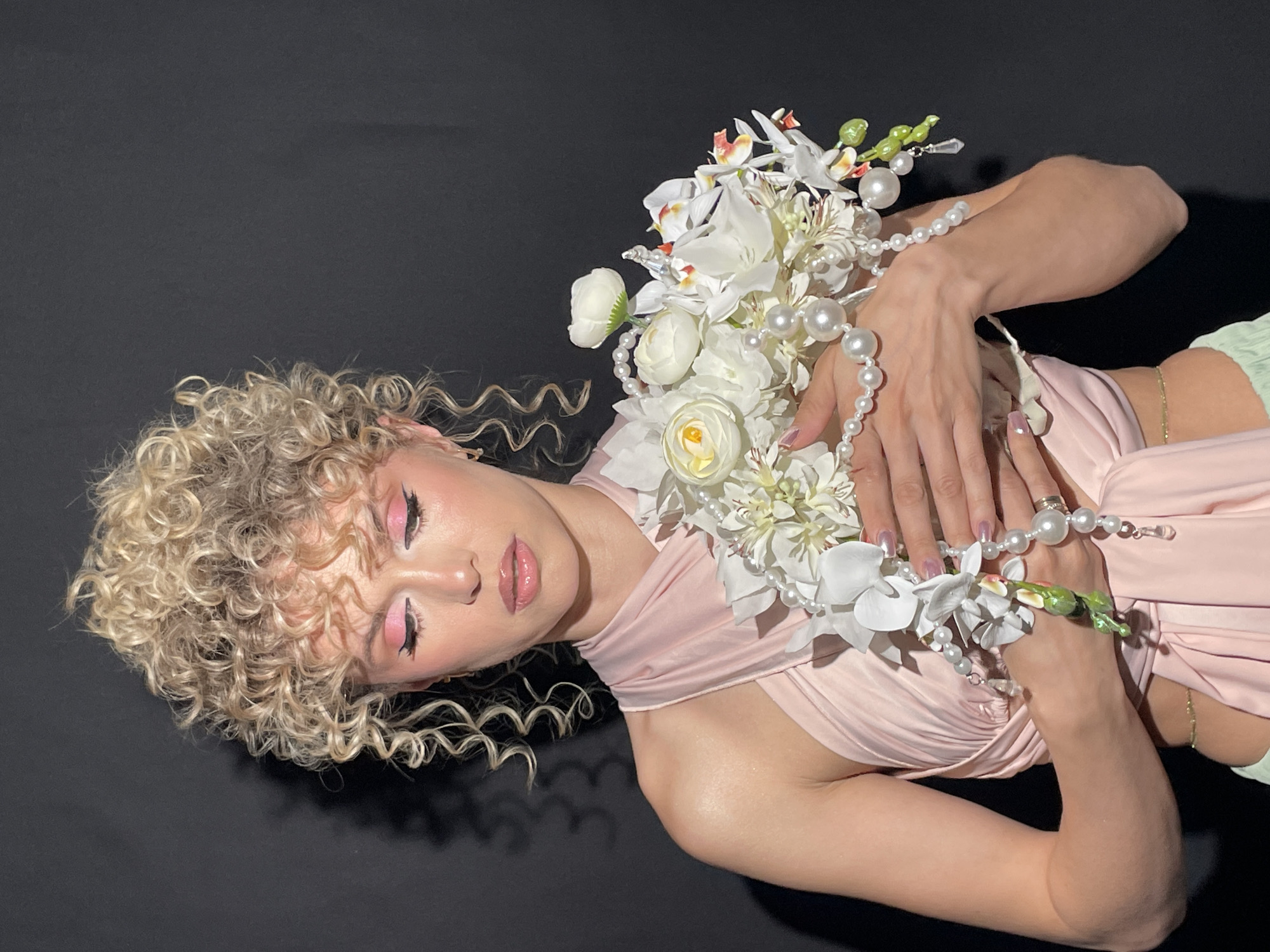 A woman with light blonde curly hair lies on a black surface. She is wearing a pale pink dress and holding a bouquet of white flowers with pearl accents. She has pink eyeshadow and glossy lips. The location is not identifiable as Kazan, Russia based on the image.