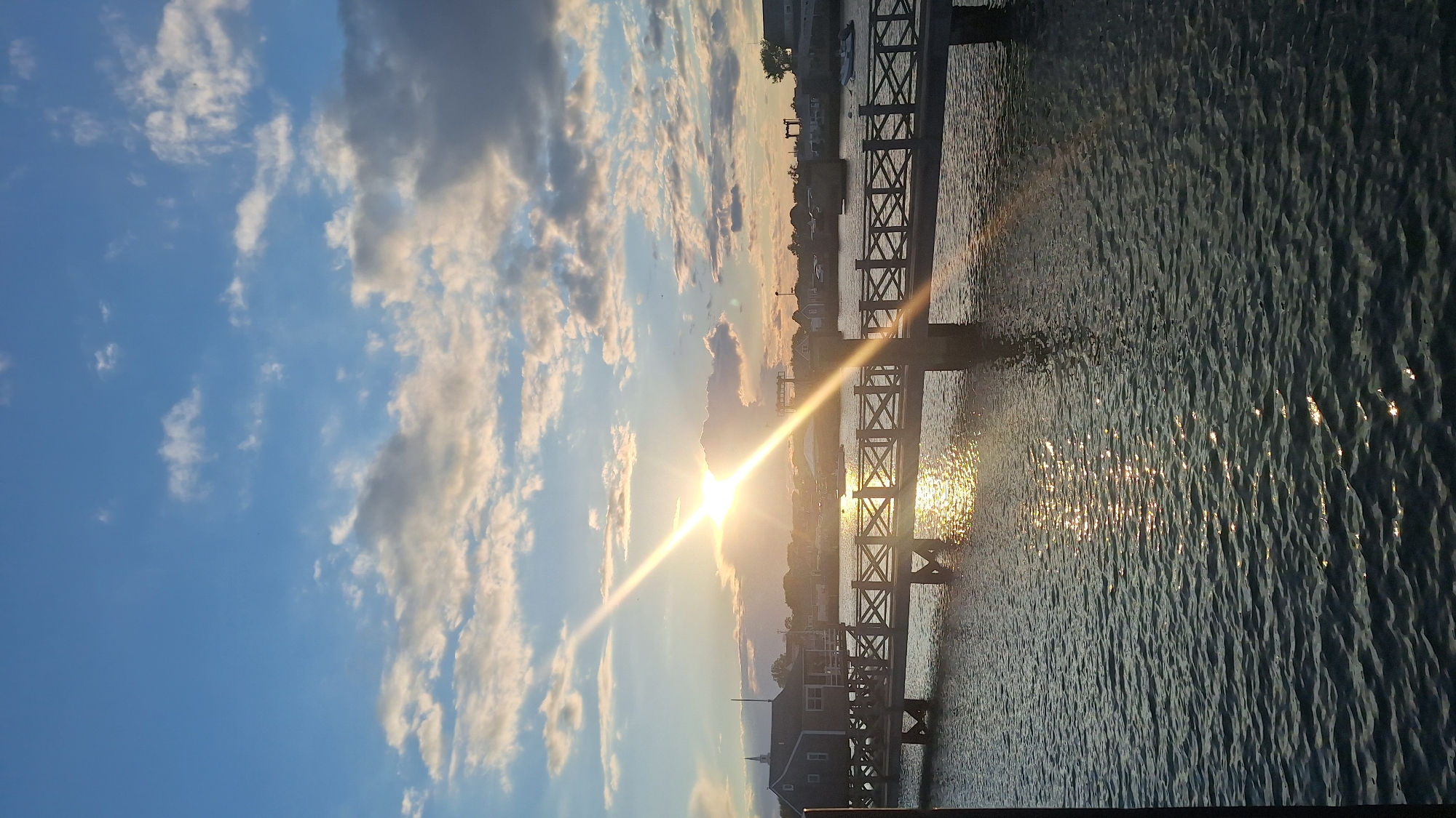This is a vertical, eye-level shot taken from a waterfront perspective, looking across a body of water towards a bridge and a town. The sun is setting on the right side of the frame, casting a strong lens flare across the image and creating bright reflections on the water. The sky is a clear blue with scattered white and gray clouds, some of which are illuminated by the setting sun.
In the foreground, the water is rippling, with the sun's reflections creating shimmering patterns on the surface. A dark, possibly wooden, pillar is partially visible on the far left, suggesting this viewpoint might be from a dock or pier.
A pedestrian bridge with a distinctive crosshatch railing spans the water from the left to the right. The bridge is supported by sturdy wooden pilings. On the left side of the bridge, a dark wooden building with a steeple, possibly a historical structure like a church or meeting house, stands on stilts over the water.
Beyond the bridge and the building, the town is visible. There are several buildings with sloped roofs, a few boats moored in the water, and some vegetation. The lighting suggests it is late afternoon or early evening. The location appears to be a harbor or a coastal town, fitting the description of Nantucket. The overall atmosphere is serene and picturesque, characterized by the warm glow of the sunset.