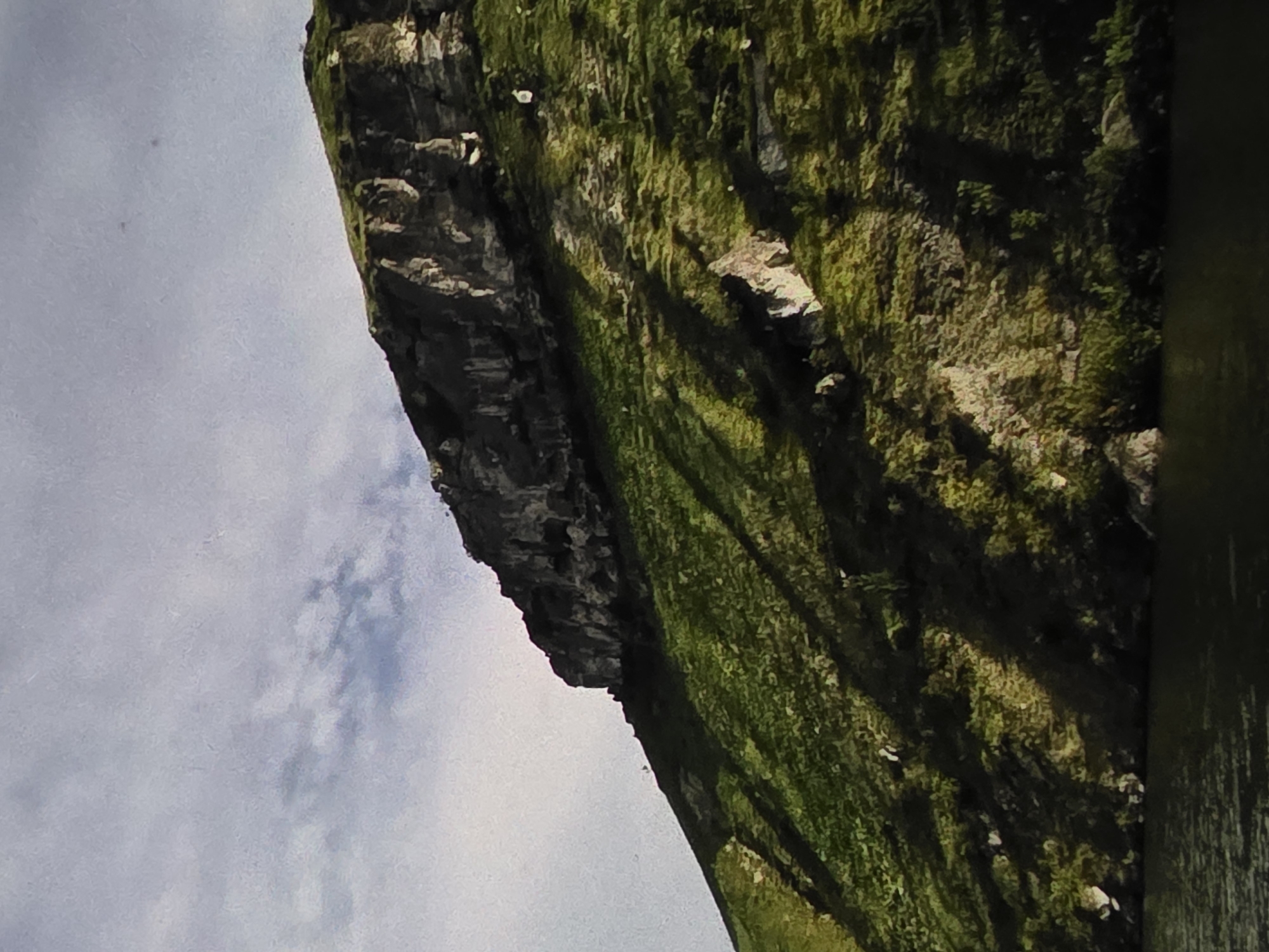 The image shows a steep, grassy mountainside rising from a dark body of water. The mountain has a rugged, rocky summit. Shadows are cast across the grassy slope, indicating it is daytime and the sun is at an angle. The sky above is partly cloudy and appears overcast. The scene is natural and undeveloped, with no signs of human structures or activity. The provided information about the location in Dubno, Ukraine, is not visually verifiable from the image itself.