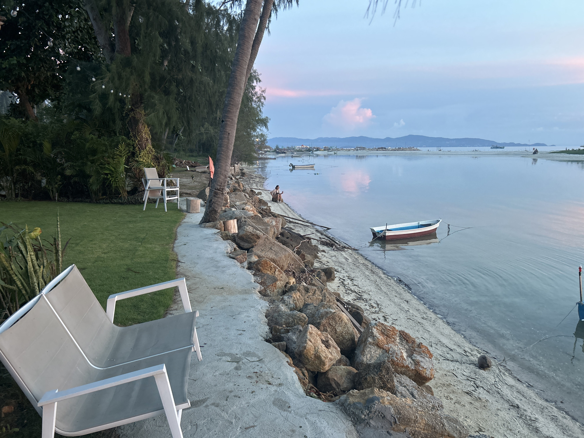 The image depicts a serene coastal scene at dusk in ตำบลเกาะพะงัน, Thailand. The foreground features a modern, grey lounge chair placed on a concrete pathway that runs alongside a grassy area with tropical foliage and tall trees. Beyond the pathway, a rocky shoreline meets calm, reflective water. Several boats are visible on the water, including a small white and red vessel anchored closer to the shore and other vessels further in the distance. The sky is a soft blend of blue and pink hues, with distant hills forming the horizon. A lone individual is seated on the rocks near the water's edge, looking out towards the sea. String lights are visible among the trees on the left side of the image, suggesting a possible resort or residential area. The overall atmosphere is peaceful and tranquil, characteristic of a tropical evening.