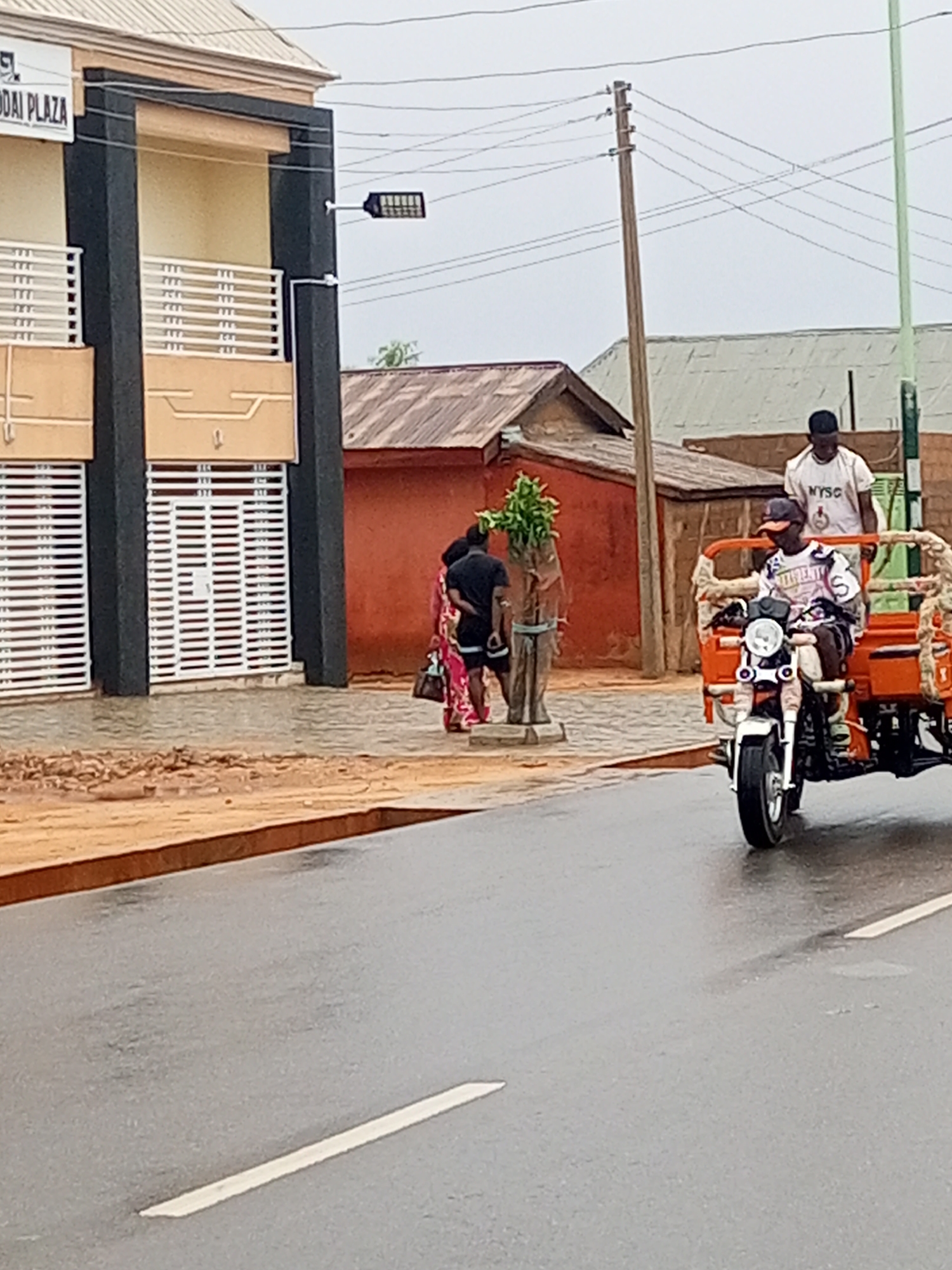 An outdoor street scene on an overcast day, likely after rainfall, shows a wet asphalt road, people, and buildings.
In the foreground, a person is driving an orange three-wheeled motorcycle, commonly known as a tricycle or "keke napep," on the wet road. The driver wears a cap and a patterned long-sleeved shirt. Another person stands in the back of the tricycle, wearing a white t-shirt with "NYSC" printed on the front.
To the left, a two-story building with dark grey pillars and light brown facades has balconies and ground-floor windows protected by white horizontal railings. A sign on the building clearly displays "UODAI PLAZA." A modern street light is mounted on one of the building's pillars.
On the sidewalk area between the building and the road, two individuals are standing. One person, dressed in a colorful patterned dress, is carrying a handbag. The other, wearing a dark top and shorts, stands near a decorative wooden post topped with green foliage. The sidewalk itself appears to be a mix of dirt and gravel.
In the background, behind the main building, several smaller structures with reddish-brown walls and corrugated metal roofs are visible. A wooden utility pole with numerous power lines stretches across the frame against the grey sky. The road features a visible white line. The overall atmosphere suggests a typical day in a bustling, possibly Nigerian, urban or semi-urban environment.
