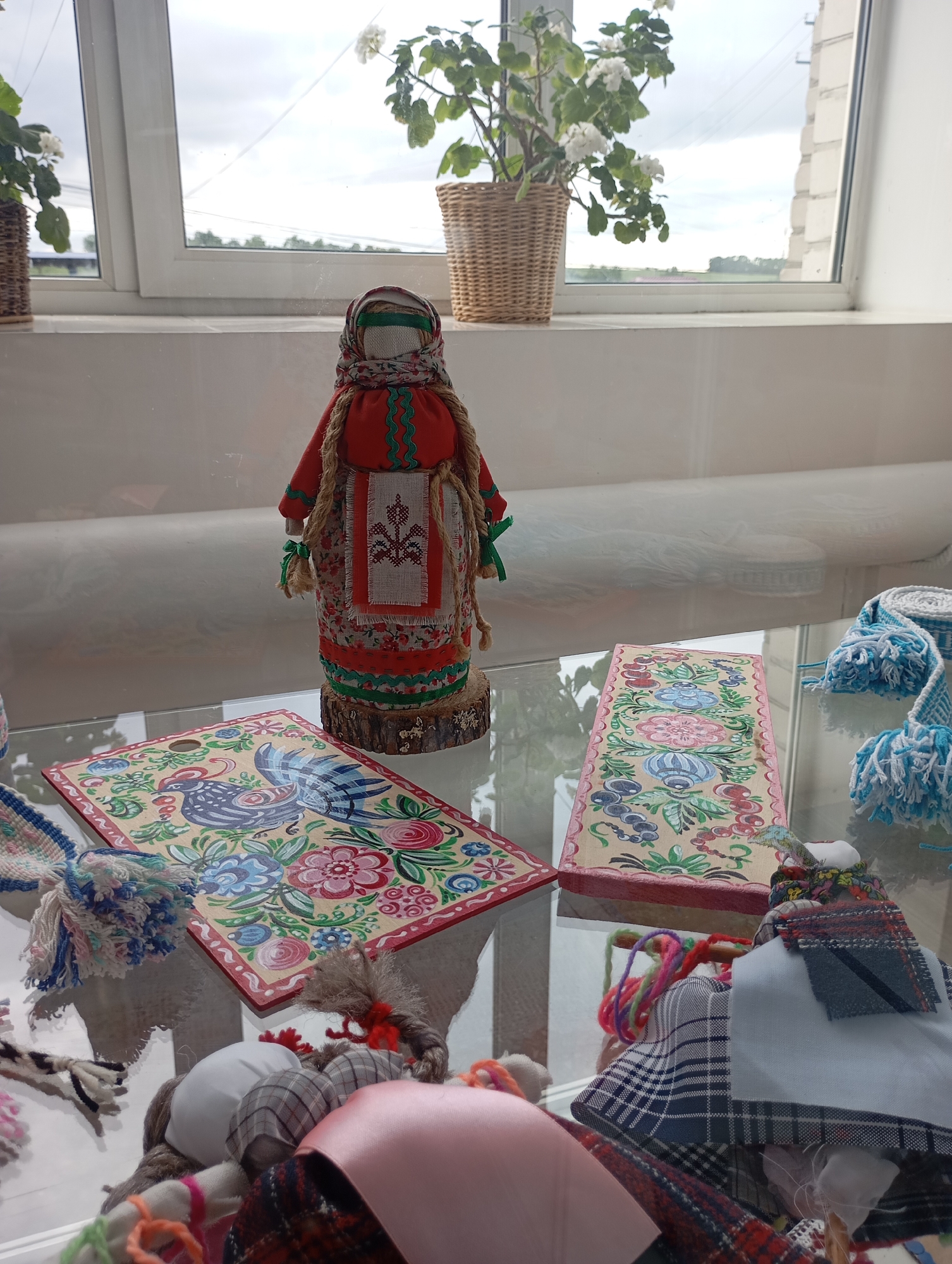 A still life shot displays a glass shelf filled with handcrafted items. A doll, dressed in traditional folk costume, stands prominently in the center, next to two painted wooden plaques with vibrant floral and bird designs. Blue and white woven pieces with tassels add color, while a collection of fabric scraps and a doll head create a sense of a work in progress. In the background, a window with a flower pot offers a glimpse of the outdoors. The location is unidentified.