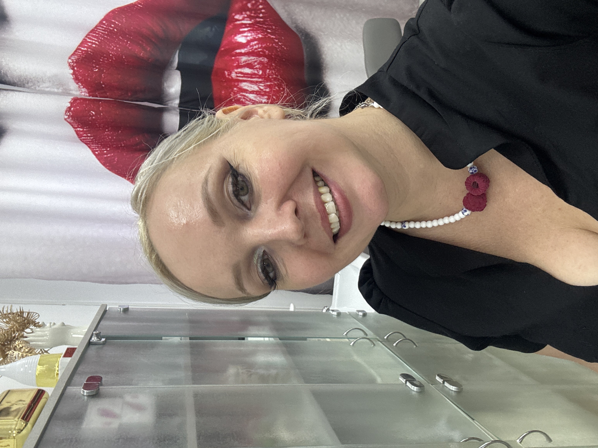 A blonde woman is taking a selfie in what appears to be a beauty salon or spa in Komsomolsk-on-Amur, Russia. She is smiling and looking at the camera. Behind her is a large, graphic print of red lips on a white background, likely a curtain or wall covering. To her left, there is a transparent cabinet with metallic handles, containing various items, some of which are obscured. She is wearing a black top and a beaded necklace with three small, raspberry-shaped adornments. Her makeup includes eyeshadow and eyeliner. The lighting suggests an indoor setting.