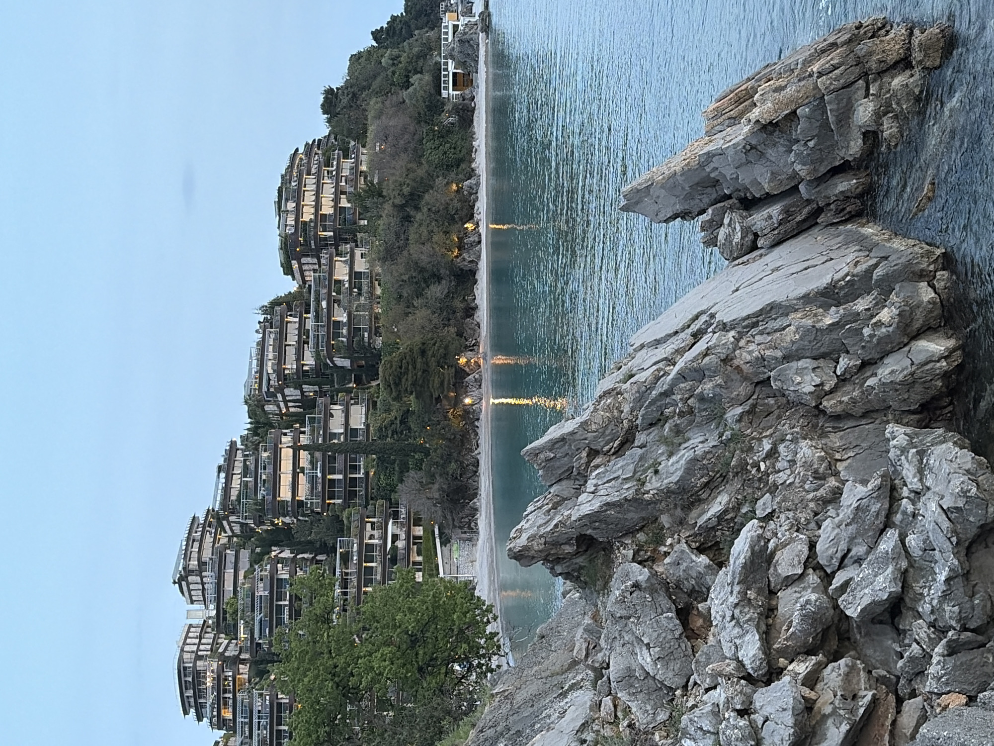 The image shows a coastal scene in Budva, Montenegro. On the left, there are tiered residential buildings with balconies, interspersed with greenery, indicating a modern architectural style. The buildings are illuminated, suggesting either evening or low light conditions.
In the foreground, there are large, gray rocks with jagged edges, typical of a rocky coastline. To the right of the buildings is a beach. Further out, the water is calm.