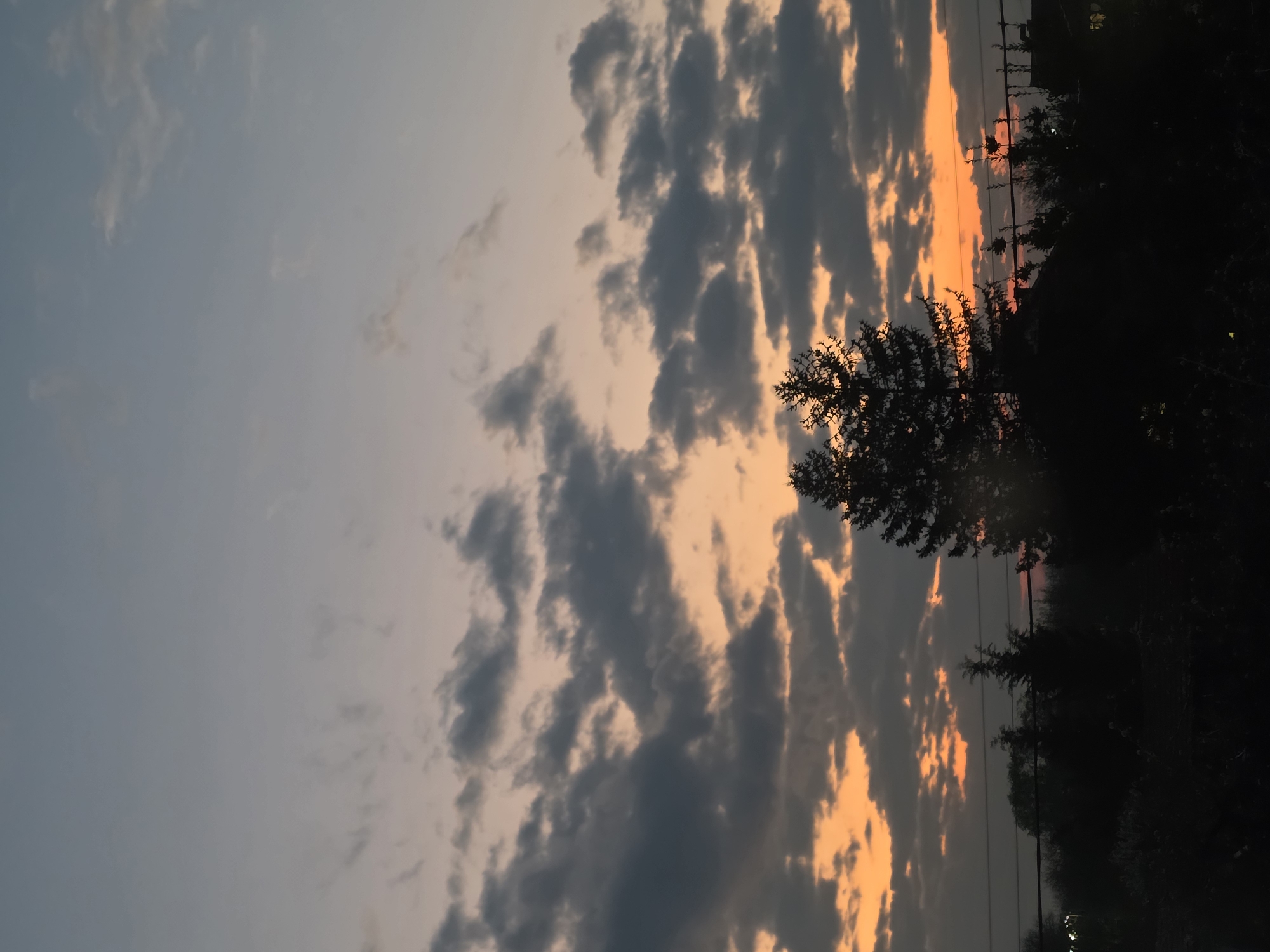 The image shows a sunset scene with a partially clouded sky over Yagodovo, Bulgaria. The clouds are illuminated with orange light from the setting sun, contrasting with darker grey areas. Silhouetted trees and vegetation are visible against the bright sky. Power lines are also silhouetted, suggesting the presence of infrastructure. The time is likely late afternoon or early evening based on the sunset. There are no people visible.