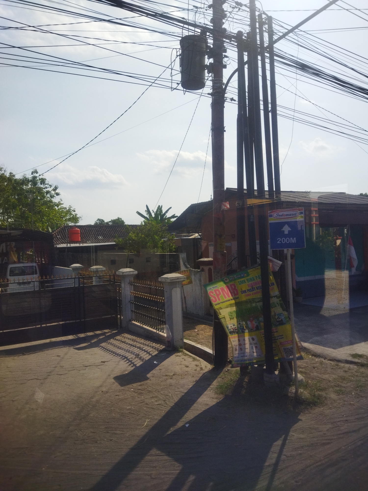 In the city of Nguter, Indonesia, a street scene unfolds under a bright sky crisscrossed by numerous electrical wires. A utility pole, adorned with a transformer and multiple conduits, stands prominently. Attached to the pole is a blue sign indicating "Indomaret" and an arrow pointing to the right with "200M" below it, suggesting a nearby Indomaret store. Leaning against the pole are two posters, one a yellow flyer with images and text, and another smaller, green flyer with "SPMB" visible. To the left of the pole, a black metal gate with ornate fencing leads into a property. Beyond the gate, a white van is partially visible, parked next to a covered structure. In the background, residential buildings with tiled roofs are nestled amongst lush green trees. An orange water tank sits atop one of the roofs. The ground in the foreground is paved with cracked concrete, and shadows cast by the utility pole and gate stretch across the scene, indicating the direction of the sun.