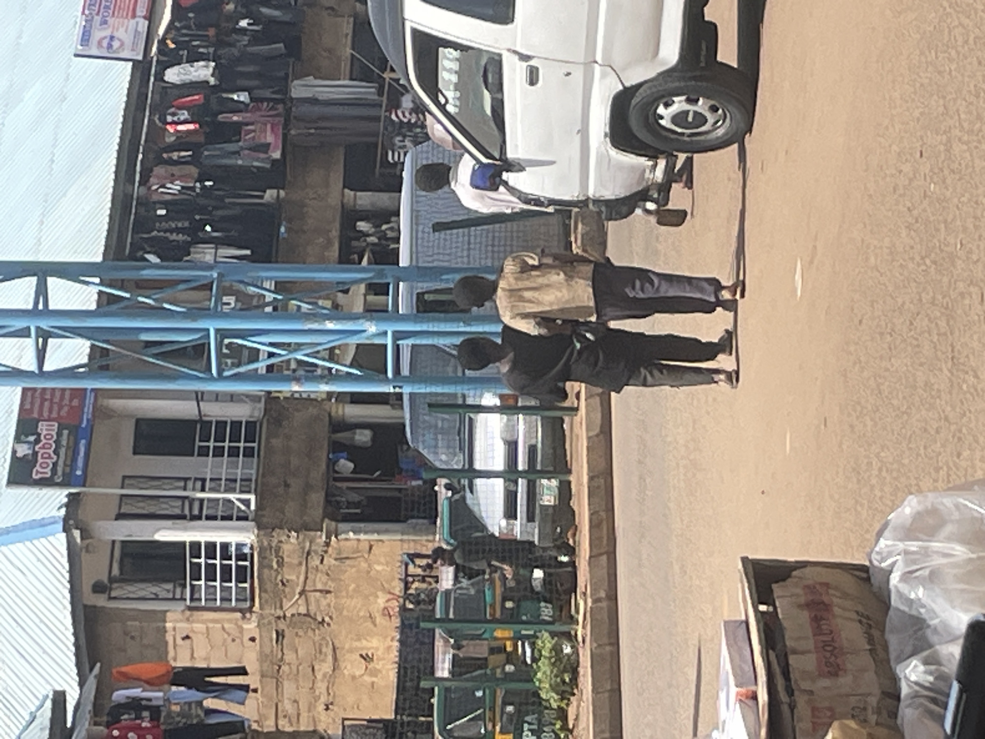 The image depicts a street scene in Bukuru, Nigeria. In the foreground, a white van is parked on the right, with its front wheel clearly visible. To the left of the van, two individuals are walking away from the camera. The person on the left is wearing a dark shirt and trousers, and the person on the right is wearing a tan shirt and dark trousers. Further to the left, several auto-rickshaws, known as "keke napeps" in Nigeria, are lined up along the curb. One of these keke napeps has the registration number "PTA 3478". Behind the keke napeps, a person is standing and looking towards the right. The background shows a commercial area with buildings housing various shops. Clothing is displayed for sale outside some of these establishments, hanging from racks. A sign with "TopBoil" and other text is visible on one of the buildings. The overall environment appears to be a busy marketplace.