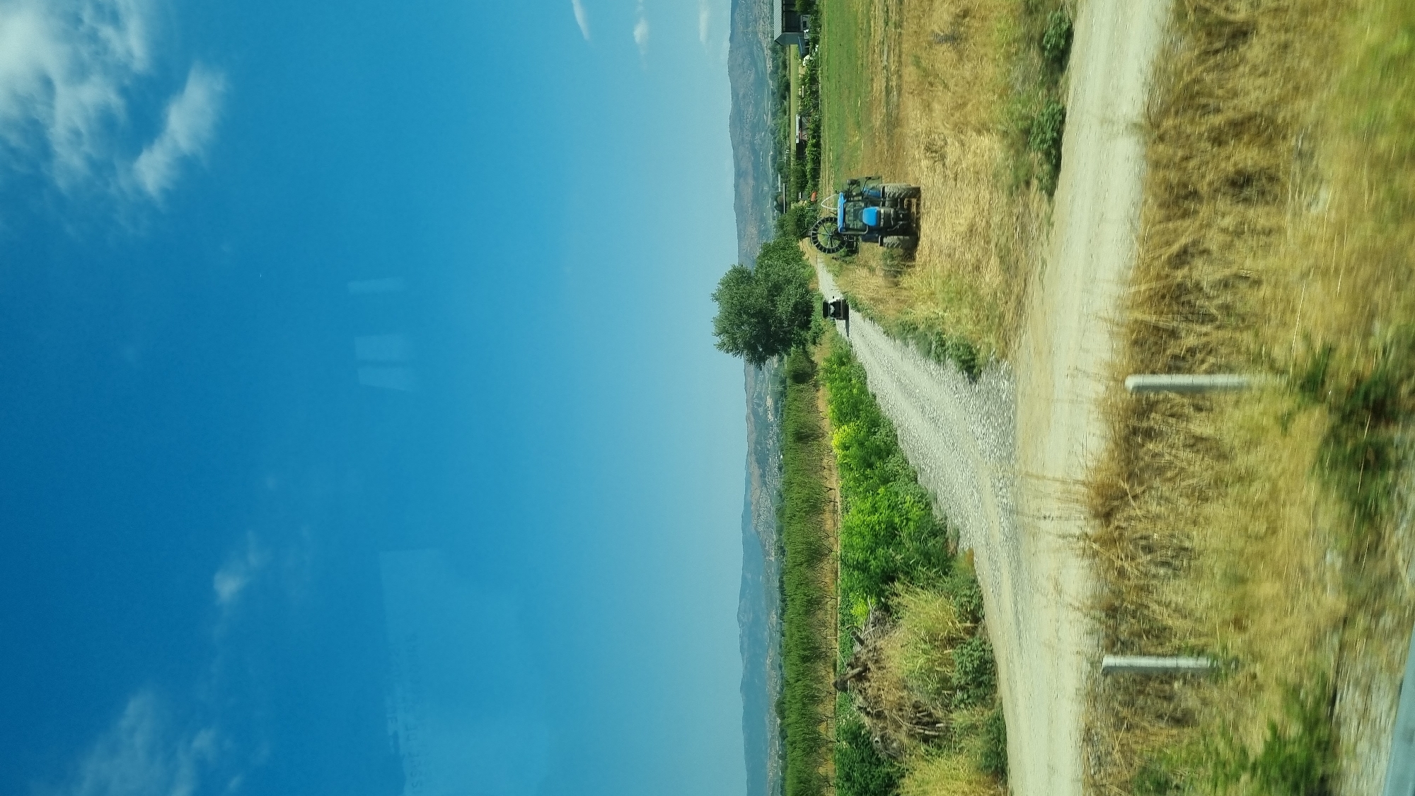 This image depicts a rural scene under a clear blue sky with scattered clouds. The foreground shows dry, yellowed grasses and two fence posts. A dirt road winds through the landscape, bordered by green vegetation and fields. Further down the road, a blue tractor is visible in a field of dry grass. In the middle ground, a dense green tree stands beside the road. Beyond the fields, a range of hills forms the distant horizon. The location is identified as Raxa, Greece. There are no people or overt actions visible in the scene. The overall impression is one of agricultural land and a peaceful, sunny day.