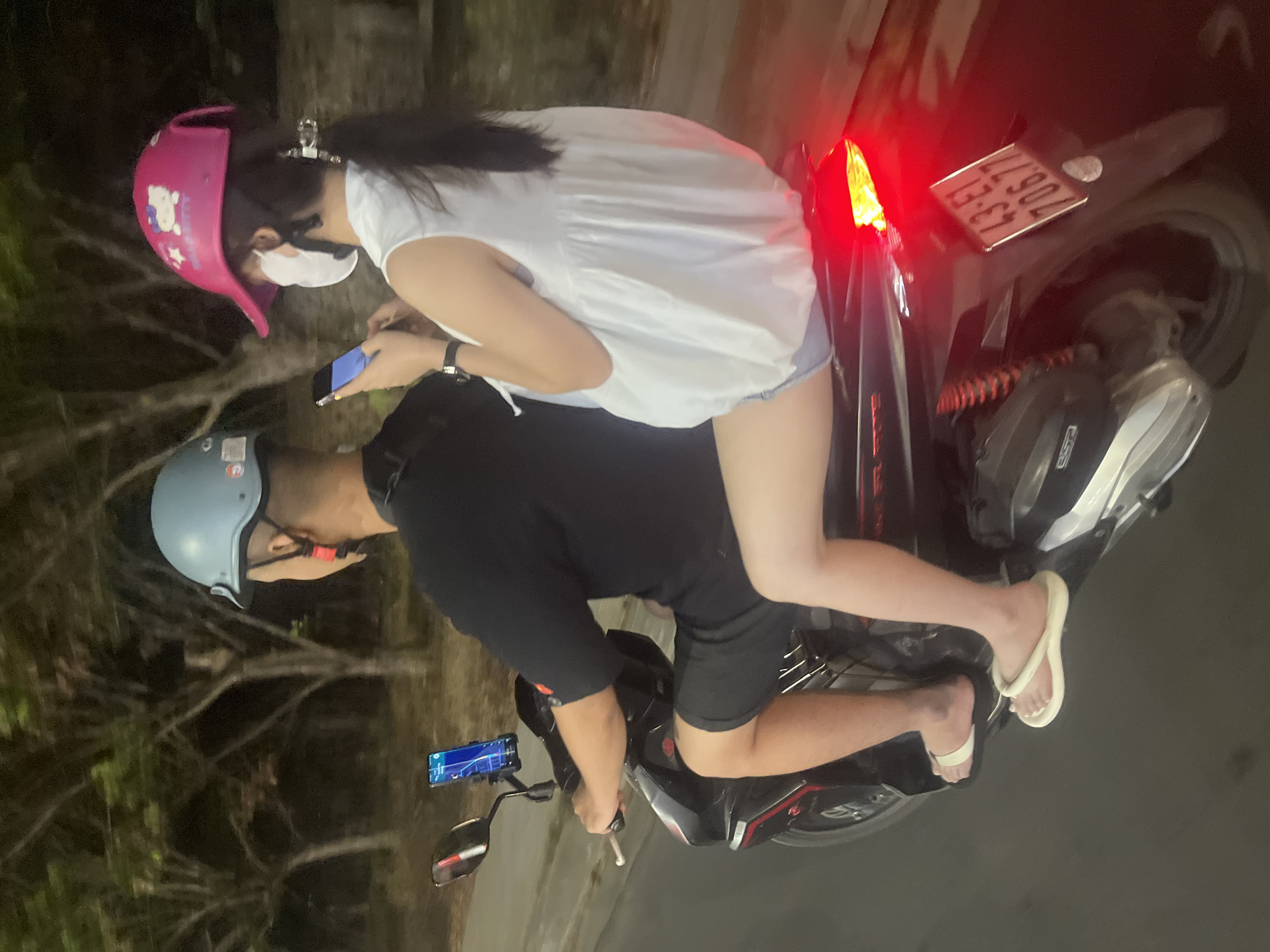 Two individuals are riding a motorcycle at night on a paved road with trees lining the background, likely within the city of Đà Nẵng, Vietnam, based on the license plate. The person in front, the driver, is wearing a light blue helmet, a black t-shirt, and black shorts. Their right hand is on the handlebar. A smartphone is mounted on the handlebar to the left of the driver, displaying a navigation app. The passenger in the rear is wearing a pink helmet with a Hello Kitty design, a white sleeveless top, and denim shorts. They are wearing a white face mask and are holding a smartphone in their hands. The motorcycle has a license plate with the characters "43-E1 706.77". The motorcycle's brake light is illuminated red. The scene appears to be captured from a slightly elevated side perspective.