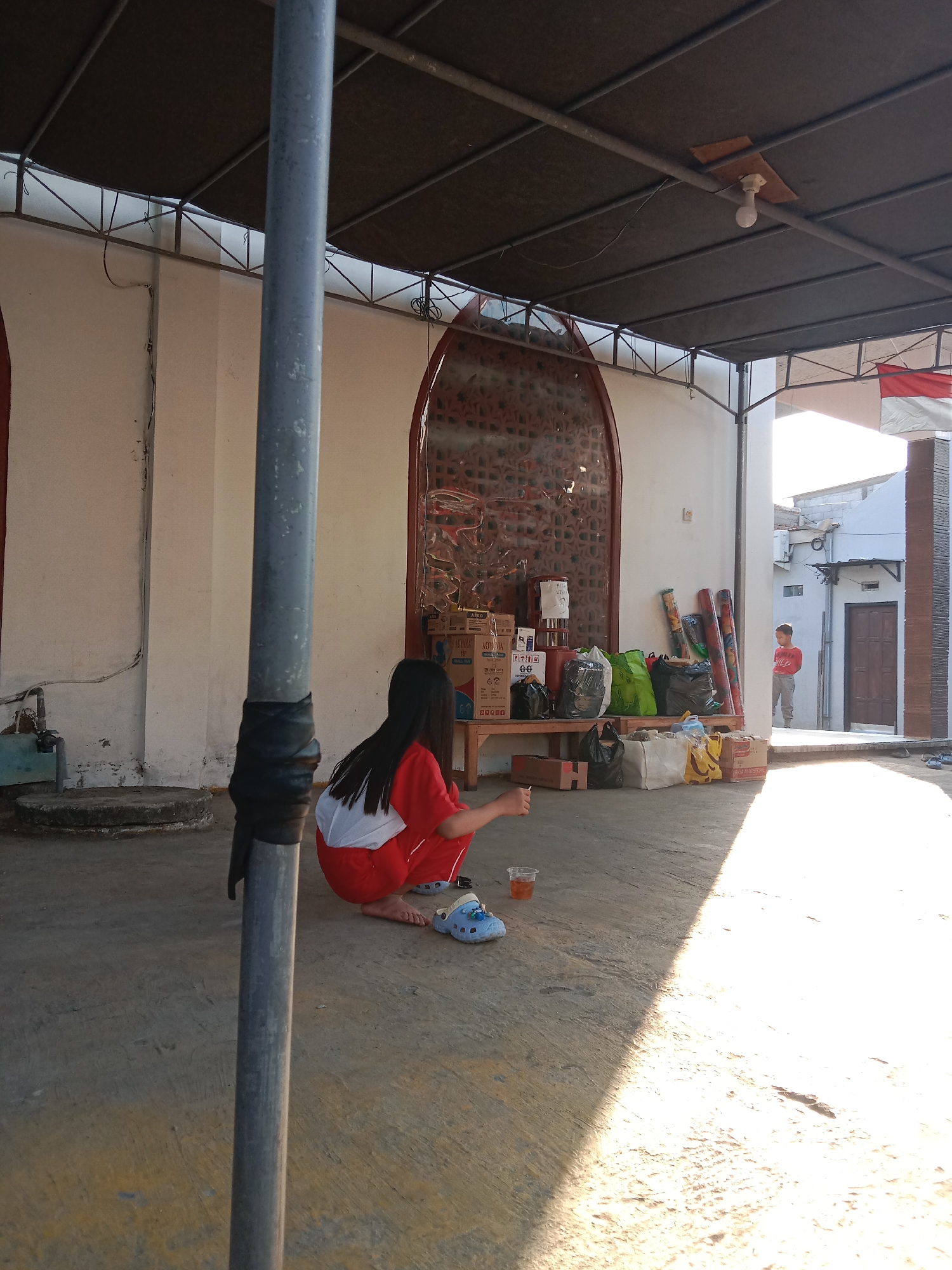 In Brangsong, Indonesia, a young girl with long dark hair, wearing a red shirt with white accents and red pants, crouches on a concrete floor. She is holding a clear plastic cup in her right hand and looking down. Beside her are blue clogs. The ground is a mix of concrete and yellowed pavement, with a strong shadow cast by an overhead structure. In the background, a wooden bench is laden with various items, including boxes, green and black plastic bags, and rolled-up carpets. A metallic archway with intricate patterns is visible on the wall behind the bench. A young boy in a red shirt and grey pants stands in a doorway further in the background. The scene is outdoors, under a dark, corrugated metal roof supported by metal beams, suggesting a sheltered outdoor area.